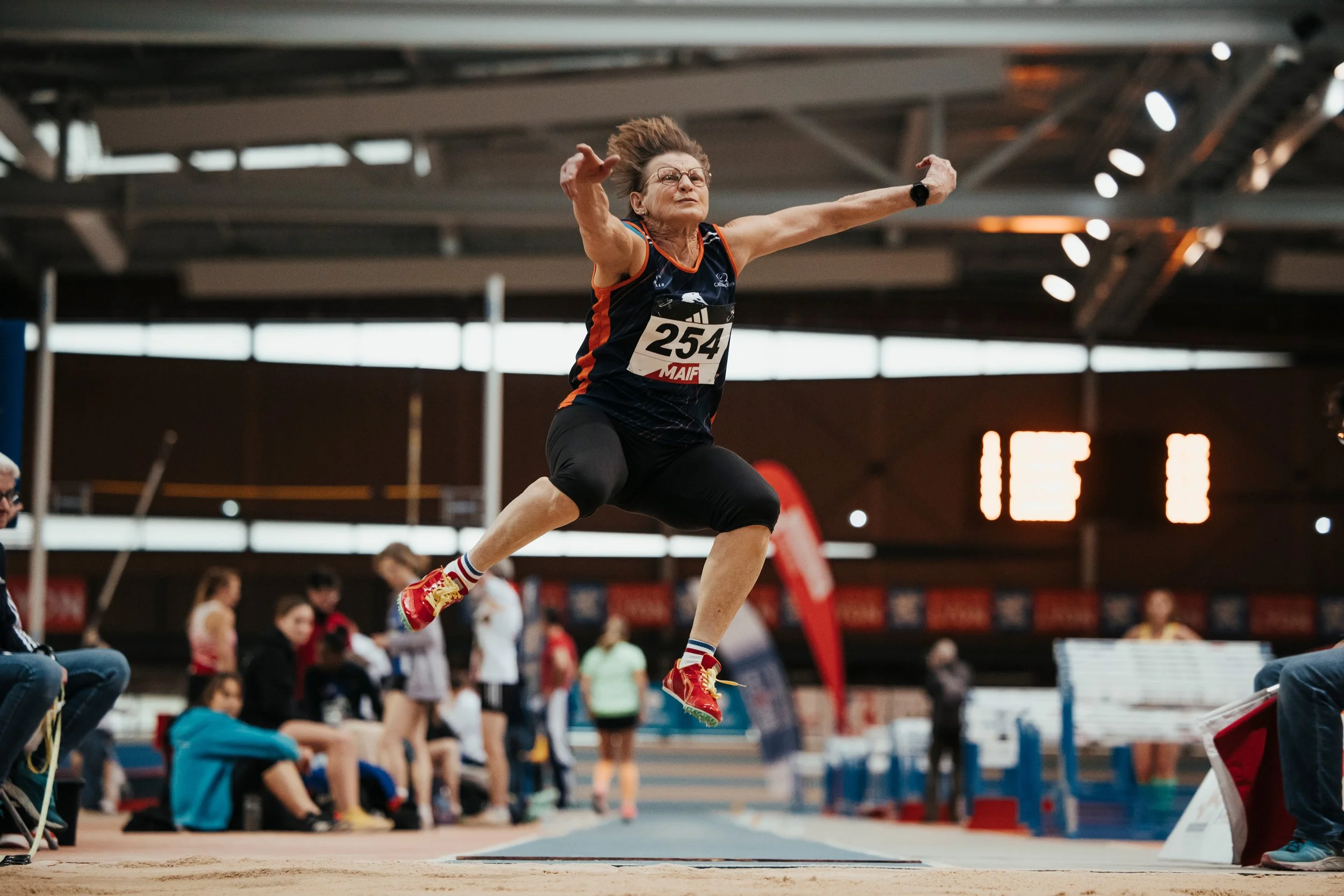 Une athlète âgée en train de sauter lors d'une compétition en salle FFA, portant un maillot bleu avec le numéro 254.