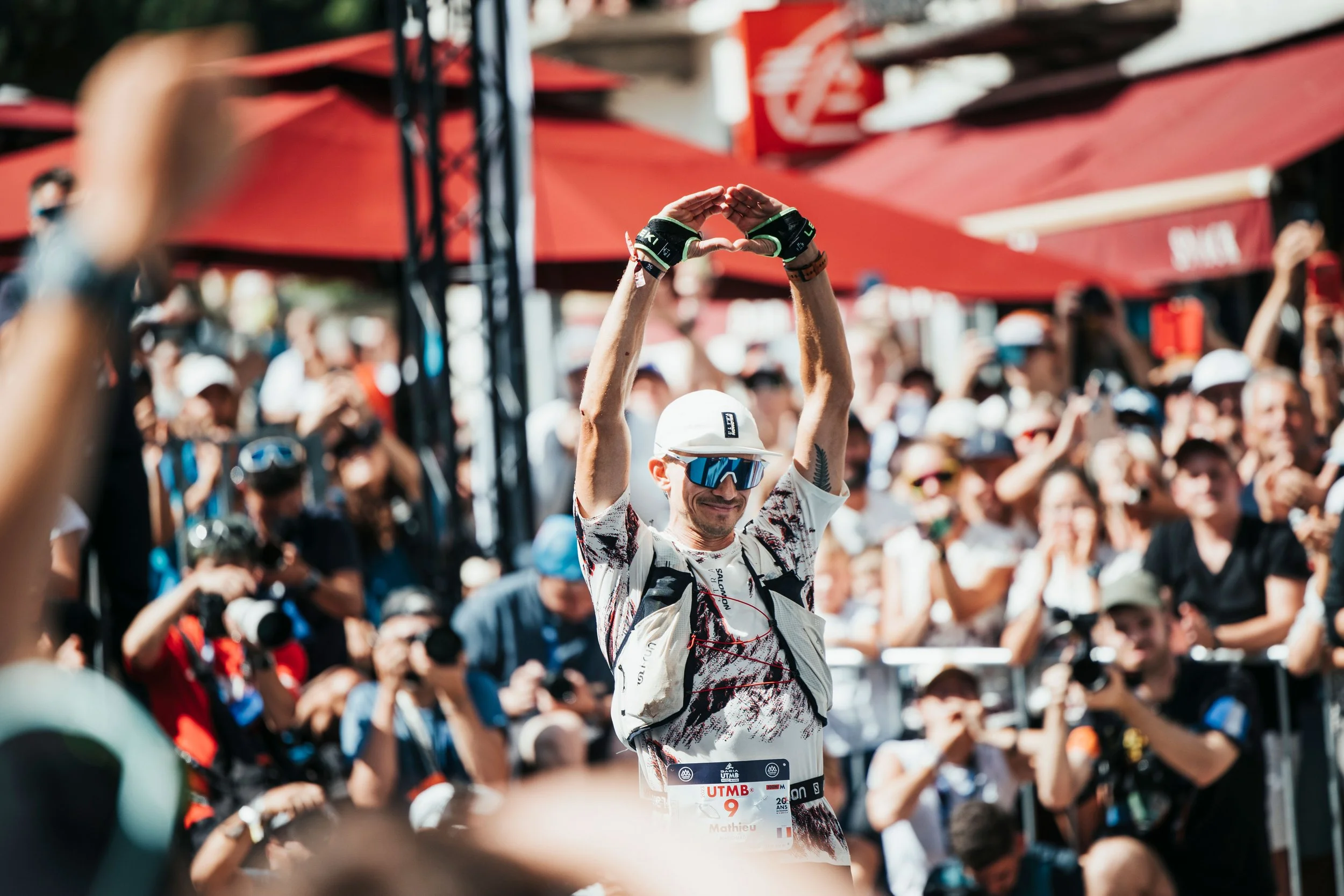 Un homme souriant, Mathieu Blanchard, portant des lunettes de soleil et un casque blanc fait un geste de cœur avec ses bras levés lors d'une course ou d'un événement avec beaucoup de spectateurs en arrière-plan.