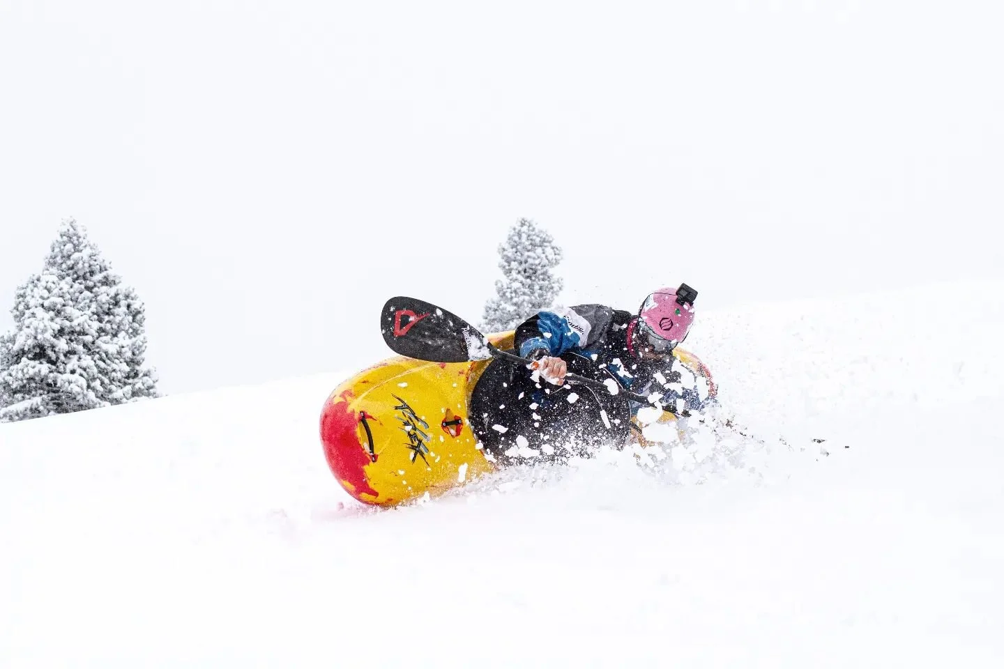 Un kayakiste, Eric Deguil en équipement de ski, portant un casque rose avec une caméra, dévale une pente enneigée avec un kayak jaune et rouge.