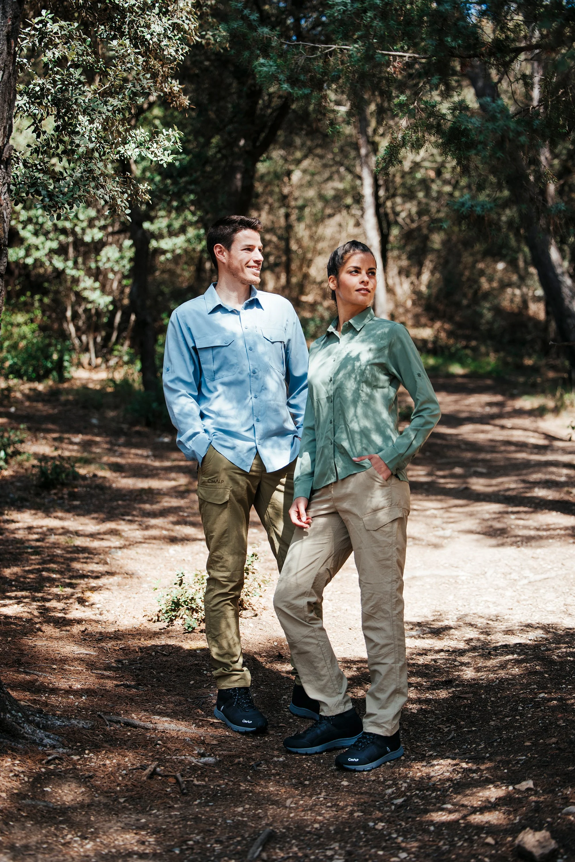 Deux personnes en tenue de randonnée dans une forêt, homme et femme, se tenant sur un sentier, avec des arbres et des feuillages en arrière-plan, en plein jour.