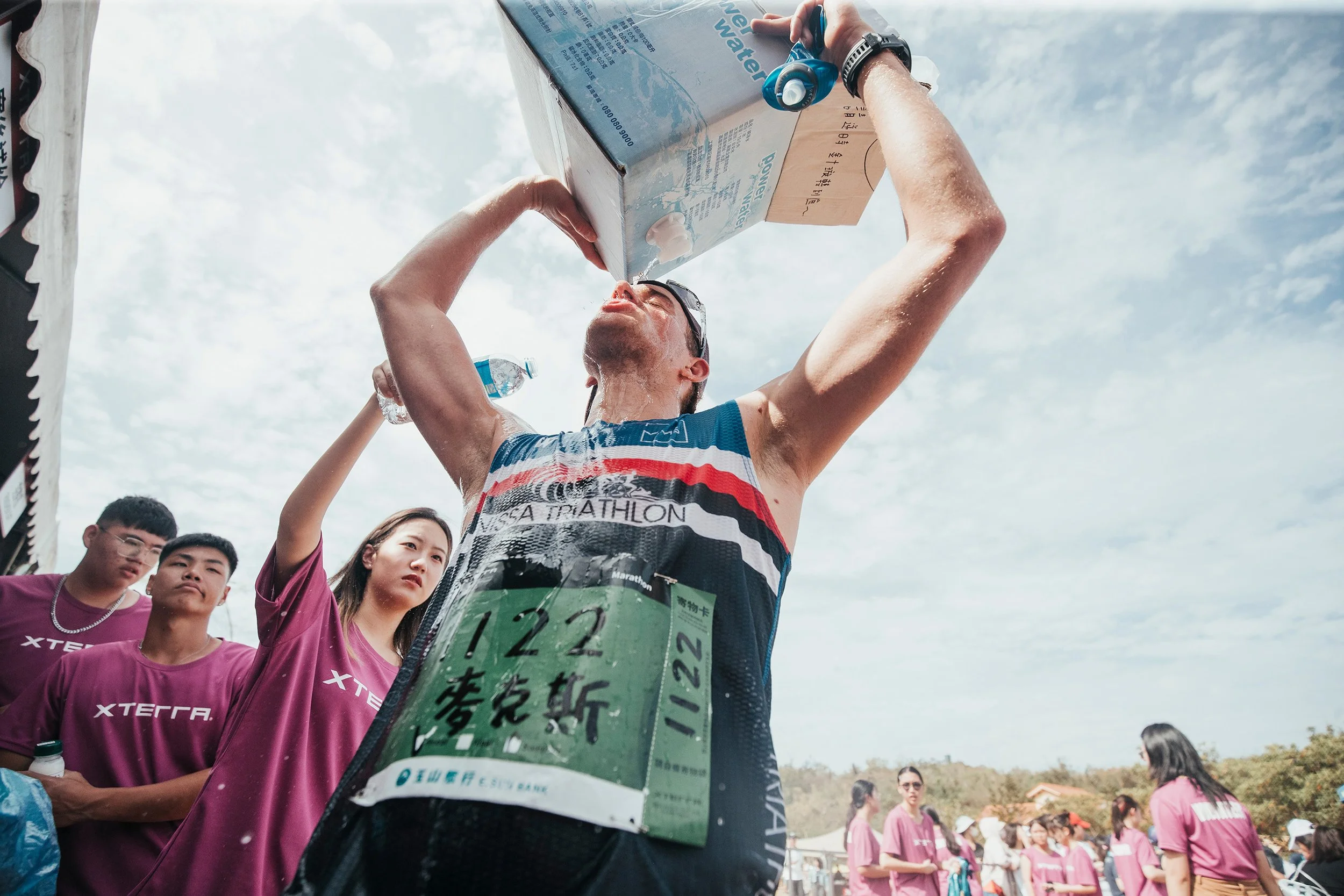 Un homme en tenue de triathlon verse de l'eau sur lui-même lors d'une course, avec un groupe de spectateurs en arrière-plan, certains portant des t-shirts roses.