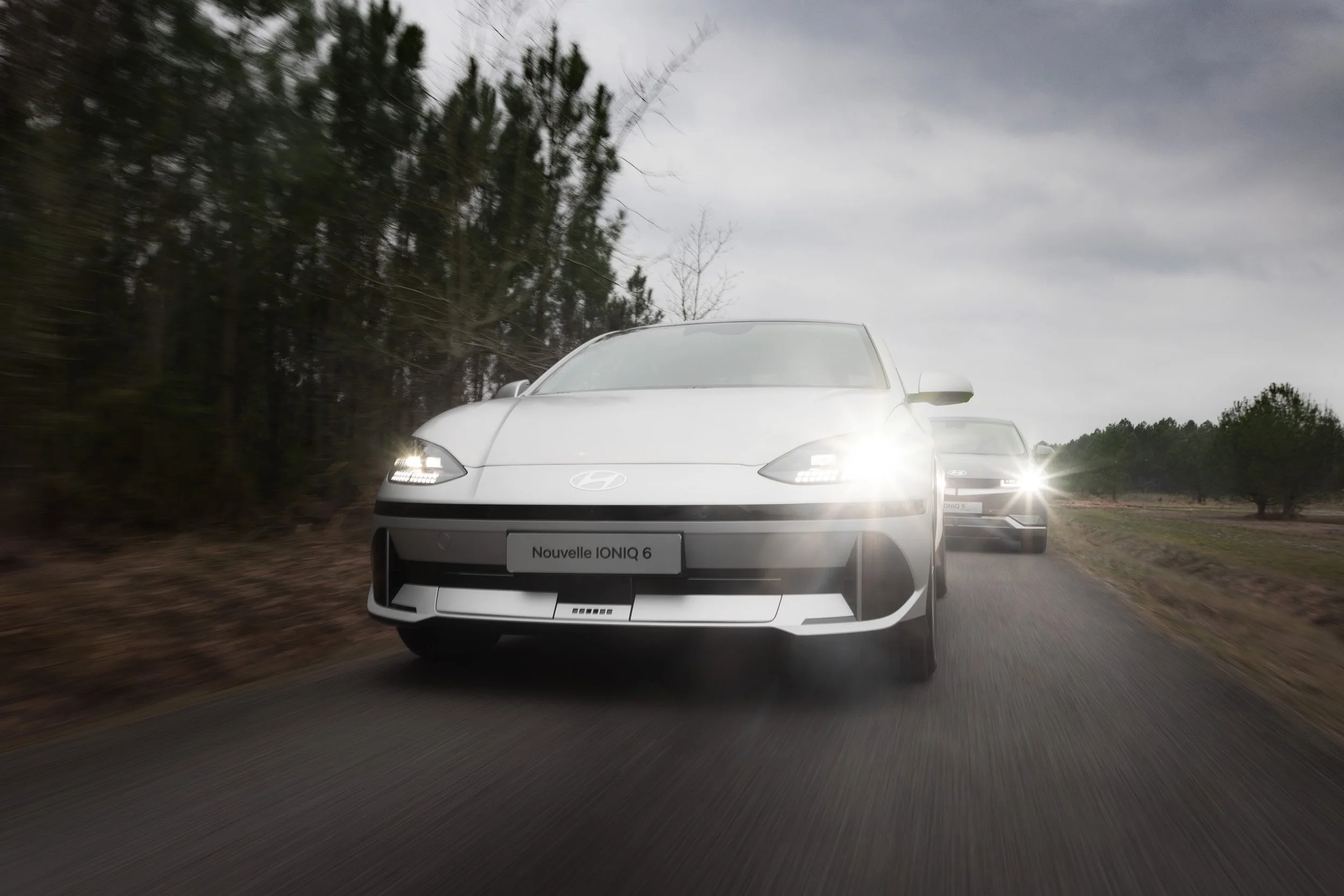 Deux voitures Hyundai, dont une nouvelle modèle IONIQ 6, en conduite sur une route de campagne par temps nuageux, avec des arbres au bord de la route.