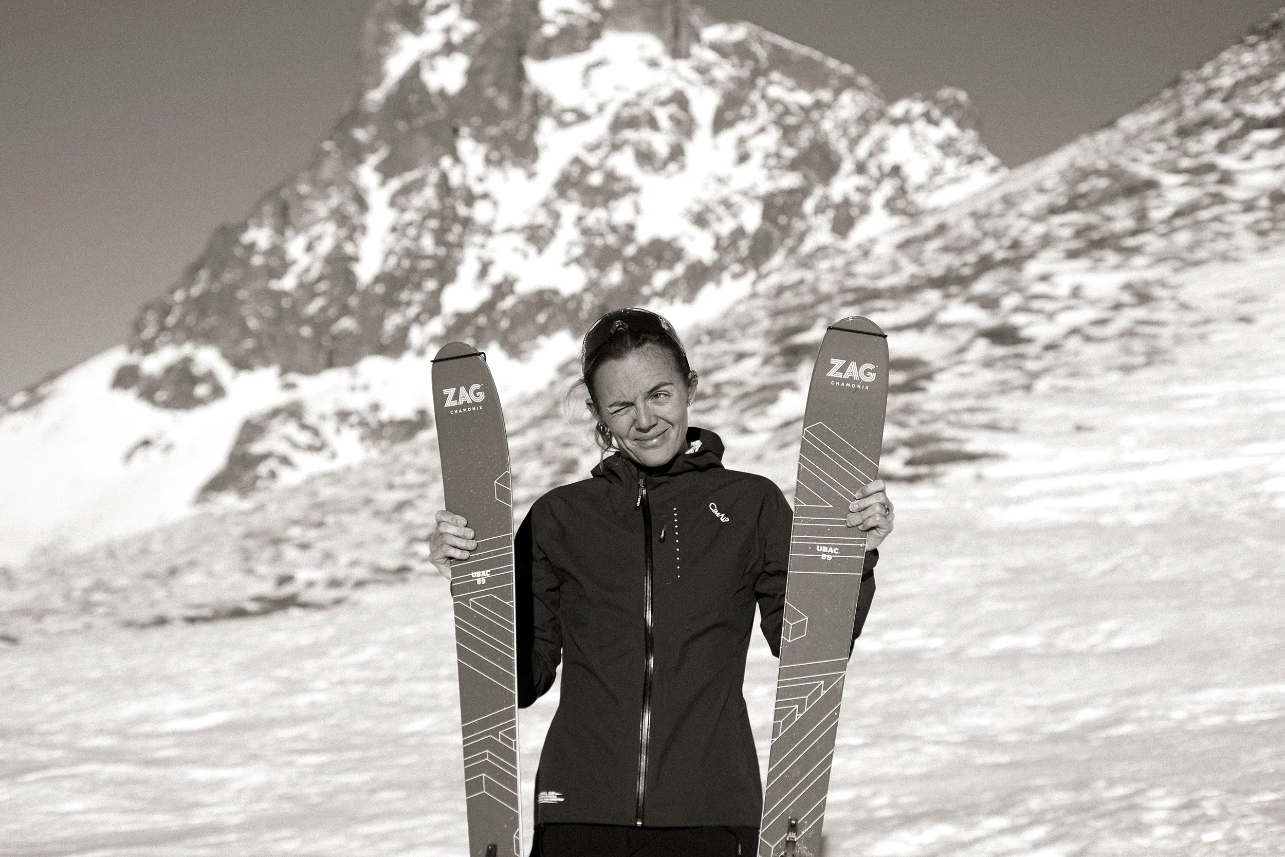 Femme souriante tenant des skis dans un paysage enneigé avec une montagne en arrière-plan.