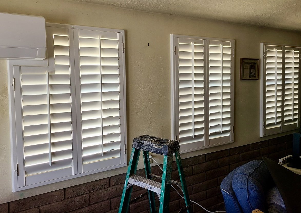 Interior view of a room with three white-framed windows with plantation shutters, a ladder, and part of a couch. An air conditioning unit is mounted on the wall.