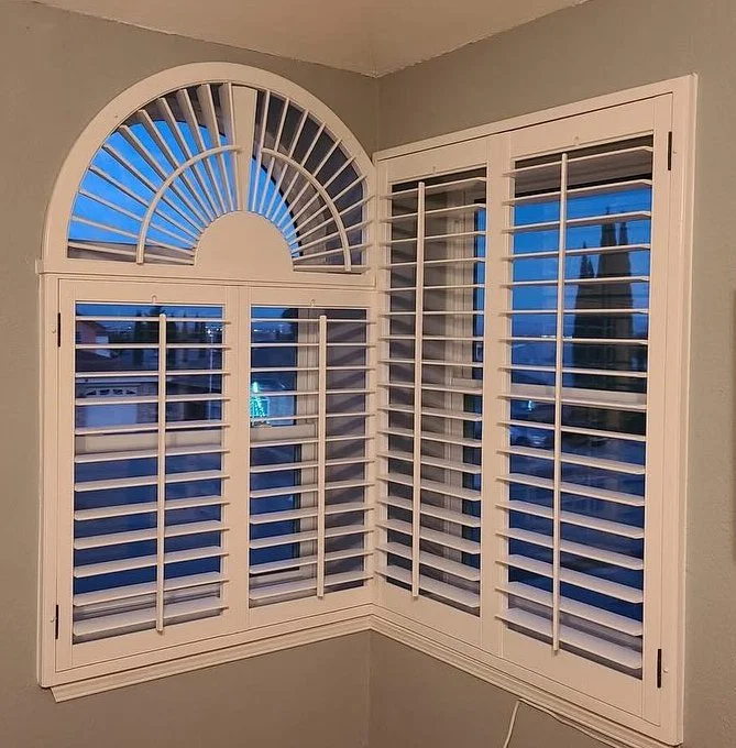 White plantation shutters on a window with an arched top section and a view of a neighborhood with houses and trees during dusk.