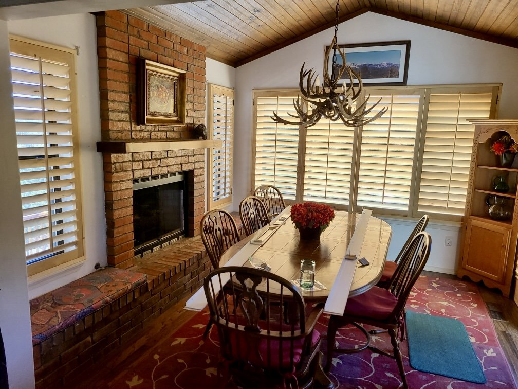 A dining room with a brick fireplace, wooden ceiling, and large windows with shutters. A wooden dining table with six chairs, a centerpiece of red flowers, and a chandelier with antler-like design. There is a window seat with a colorful cushion, a framed landscape picture above the fireplace, and a wooden cabinet with decorative items.