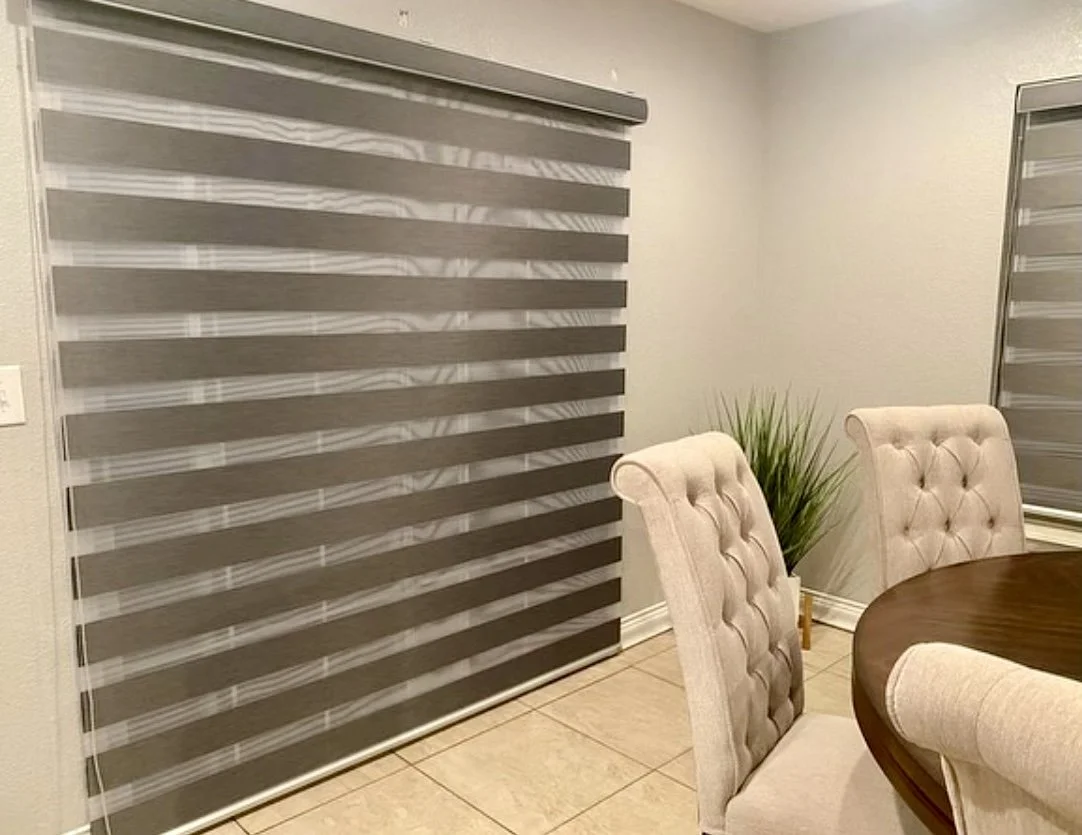 A window with zebra blinds, beige tufted chairs, a round wooden table, and a potted green plant in a corner.