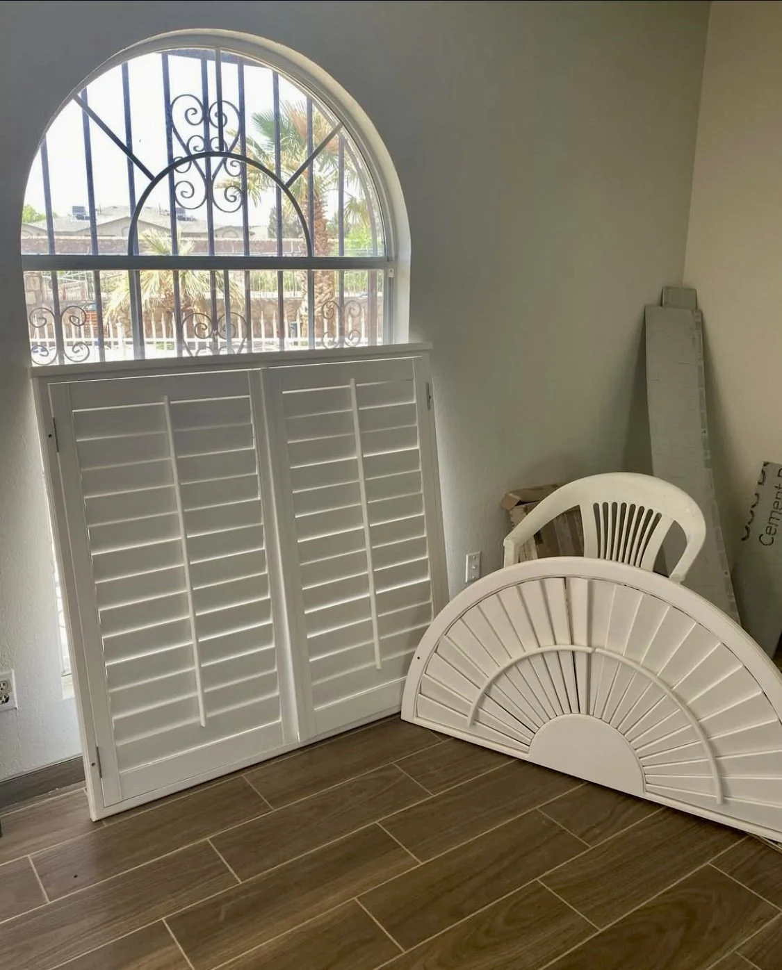 White window shutters and decorative grille, leaning against a wall, with a plastic chair and folded wood pieces nearby, on a wood-look tile floor.