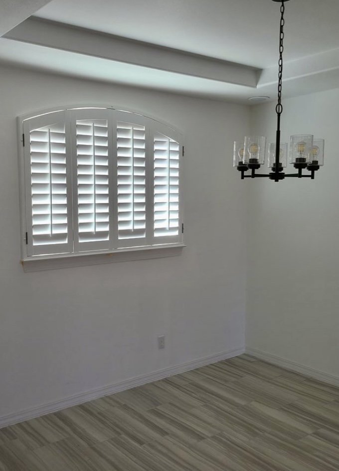 Empty room with white walls, a window with white shutters, a light-colored wooden floor, and a black chandelier hanging from the ceiling.