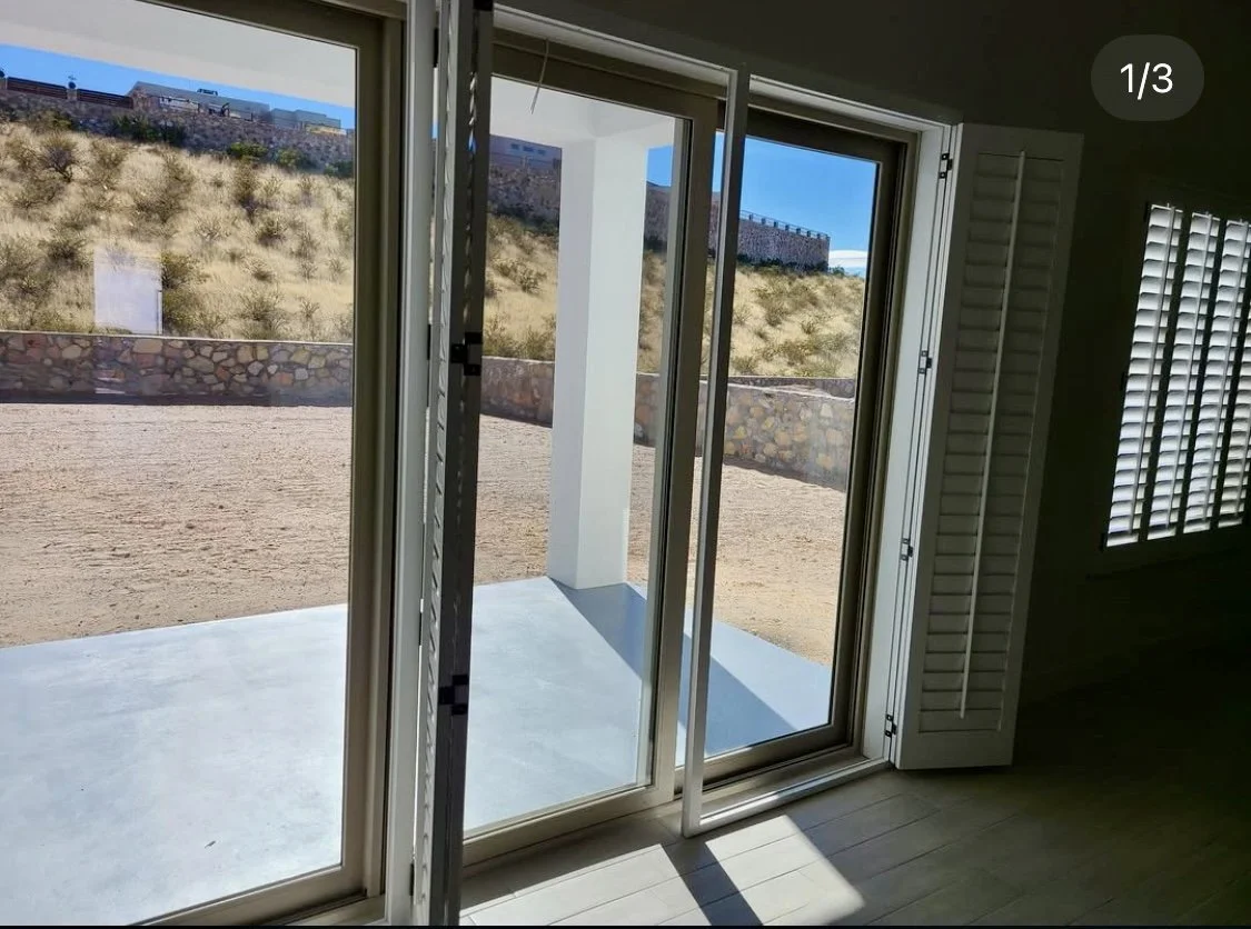 Open sliding glass door leading to a patio with a view of a hillside and a stone wall, inside a house with wooden floor and window shutters.