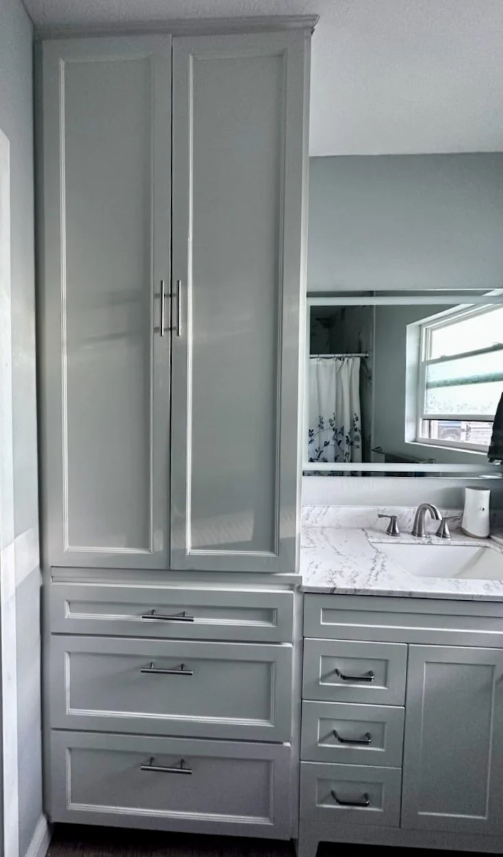 A bathroom vanity with gray cabinets, drawers, a marble countertop, and a sink with a faucet. A mirror and a window are visible in the background.