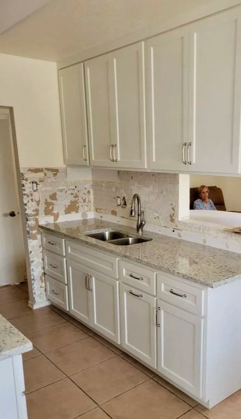 A kitchen with white cabinets and granite countertops under renovation, with peeling paint and exposed wall behind the sink, and a woman sitting in a separate room visible through an open pass-through window.