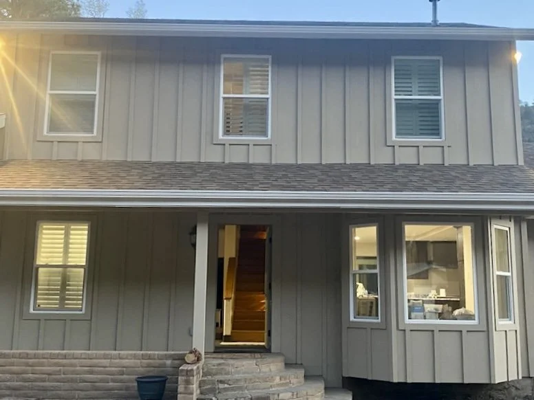 Front view of a two-story house with gray siding, three upper windows, and a porch with an open door revealing stairs inside.