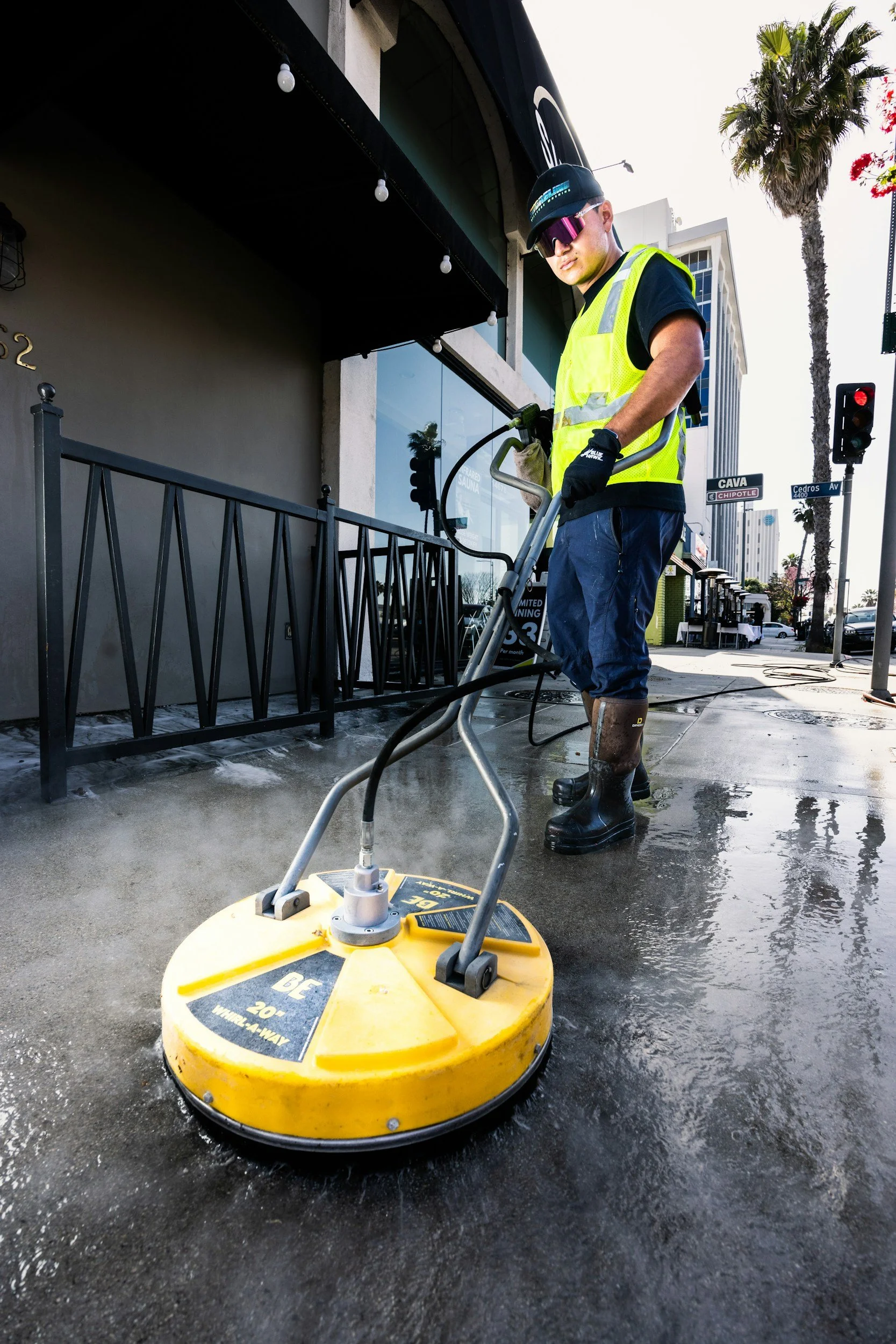 A man wearing a high-visibility vest, sunglasses, and boots uses a yellow power scrubber to clean the sidewalk outside a building on a city street.