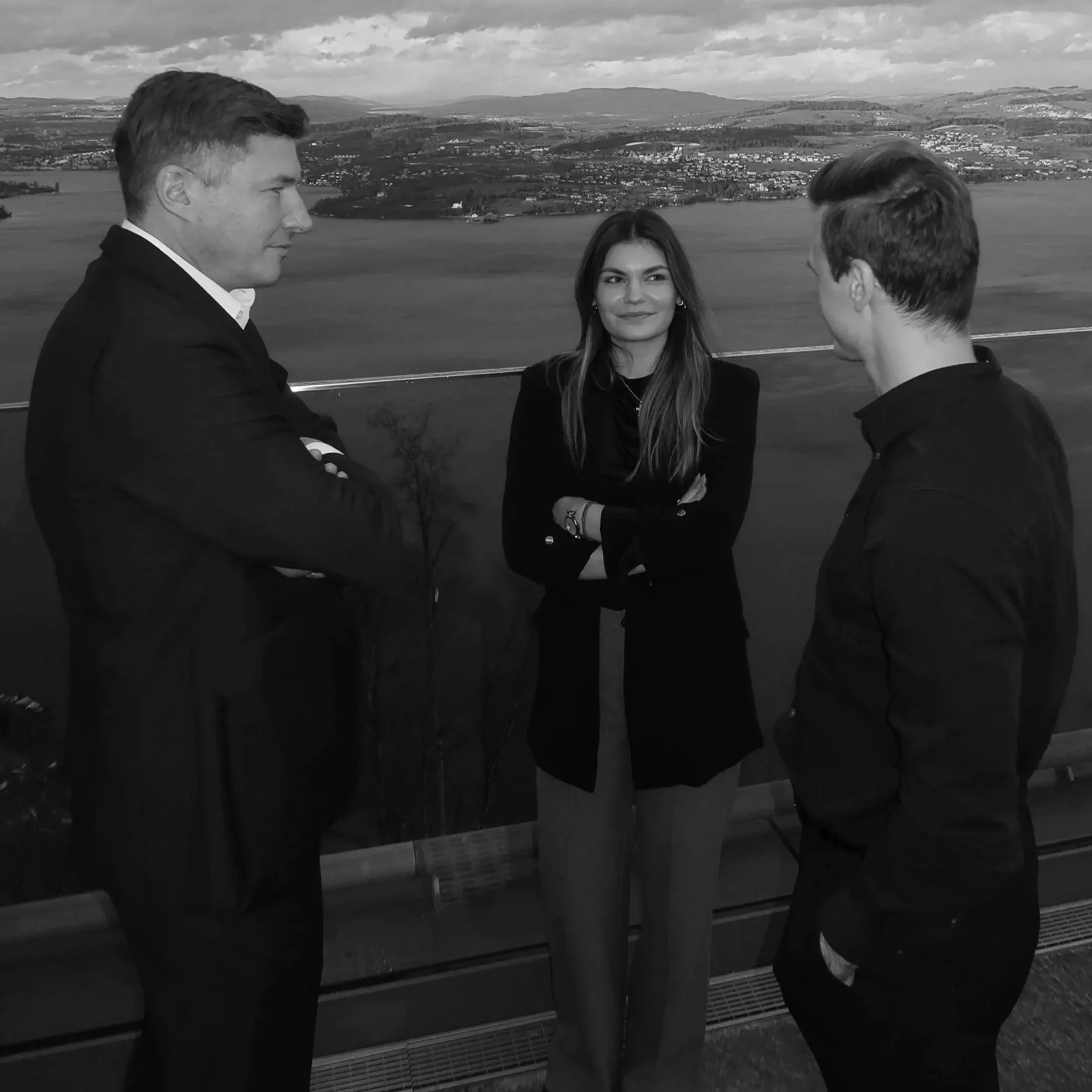 Drei Menschen stehen in einem Gespräch auf einer Balkon oder Aussichtsplattform mit Blick auf eine Landlandschaft mit See und Hügel im Hintergrund, alle tragen dunkle Kleidung.