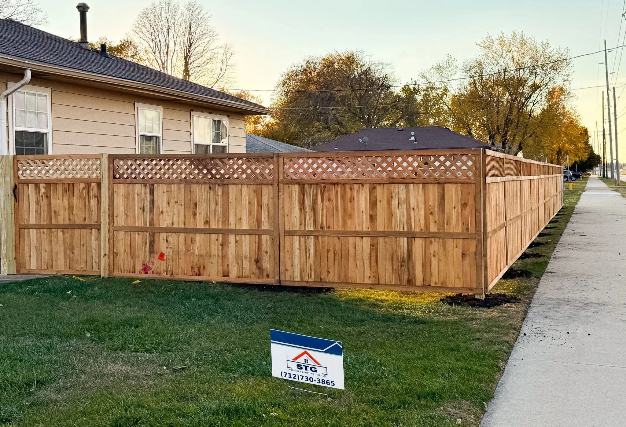 New wooden privacy fence installed in front yard of residential house; sign advertising fencing company STG with phone number.