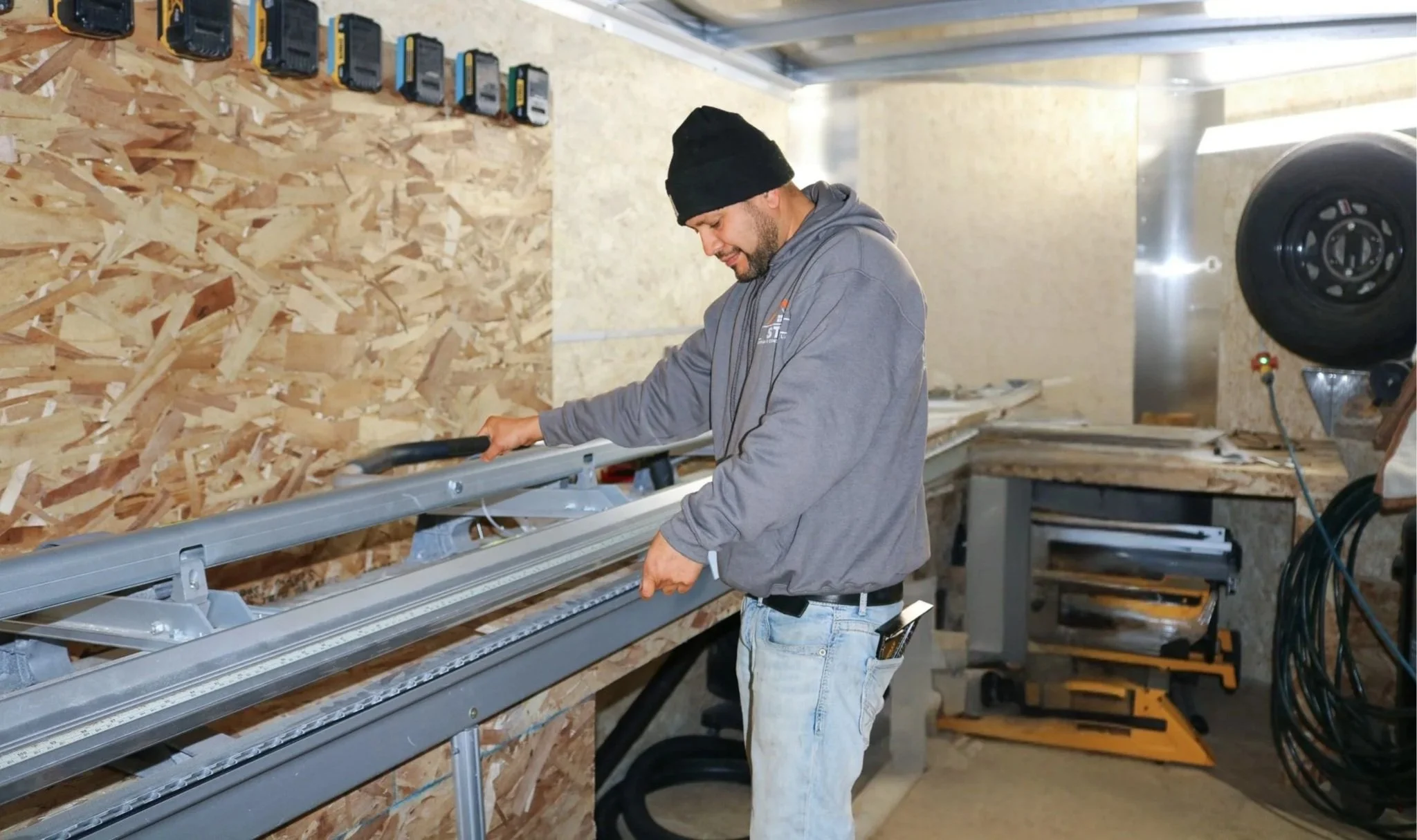 Man working with a sliding table saw in a woodworking shop, wearing a gray hoodie and black beanie.