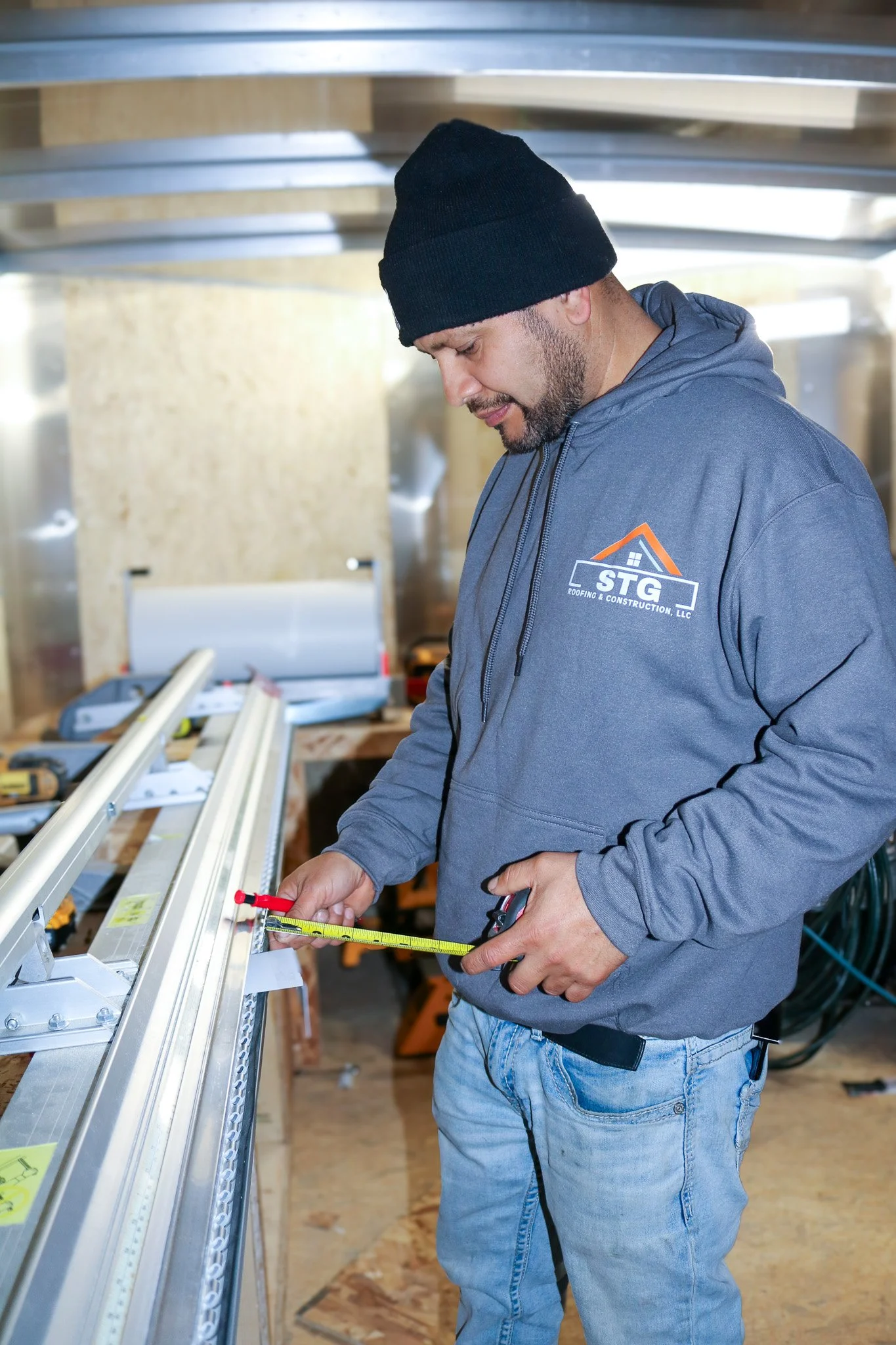 Man wearing a gray hoodie with a roofing company logo, measuring a metal frame with a tape measure inside a construction site.