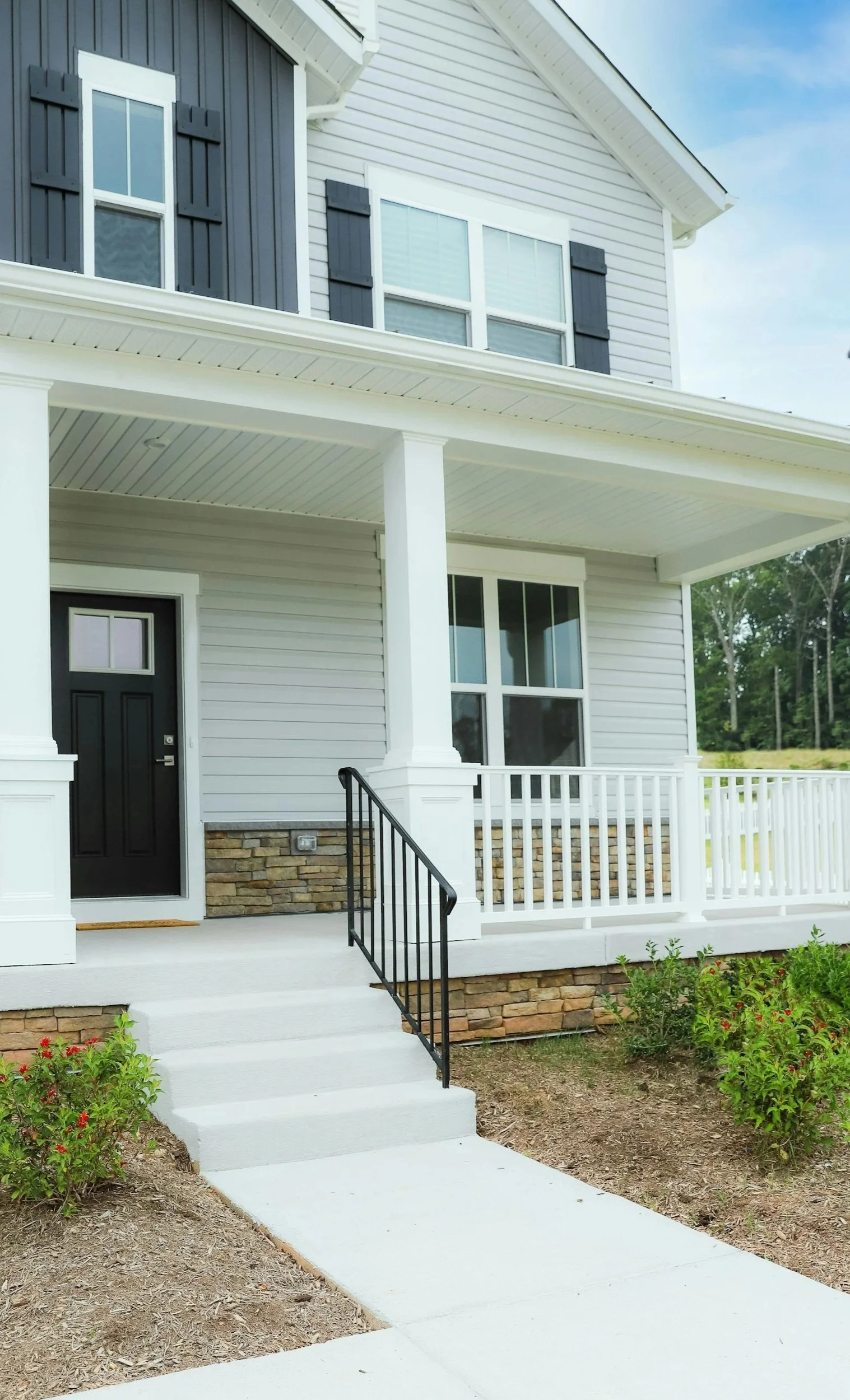 Front porch of a two-story house with gray siding, white accents, black shutters, and a black front door, with stairs and a small garden bed.