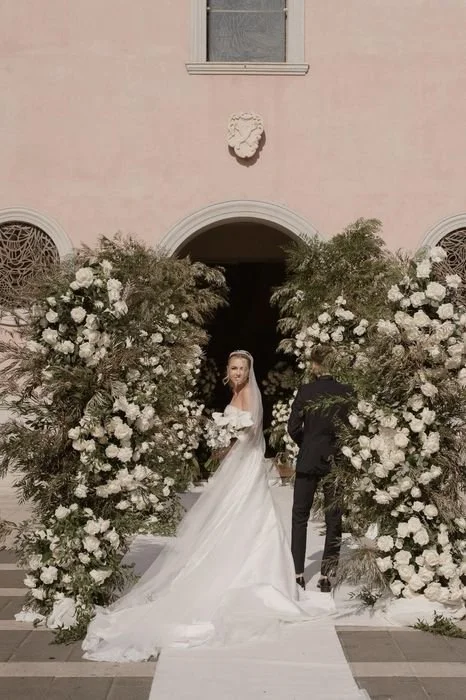 Una coppia di sposi davanti a un arco di fiori bianchi e verdi, in un matrimonio all'aperto.