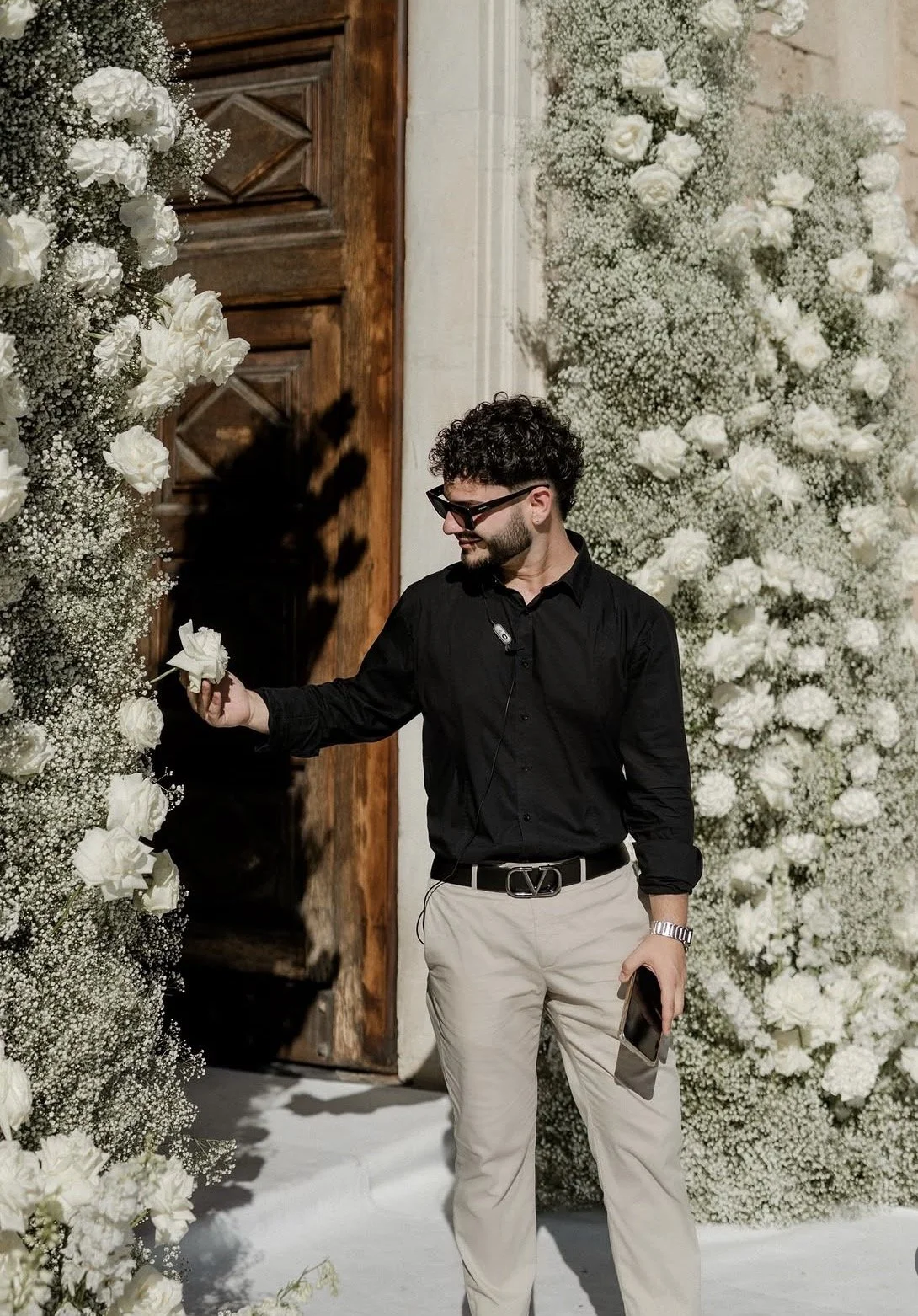 Uomo elegante con occhiali neri indossa una camicia nera e pantaloni chiari, mentre sfiora una decorazione floreale bianca in un matrimonio o evento decorato con fiori bianchi.