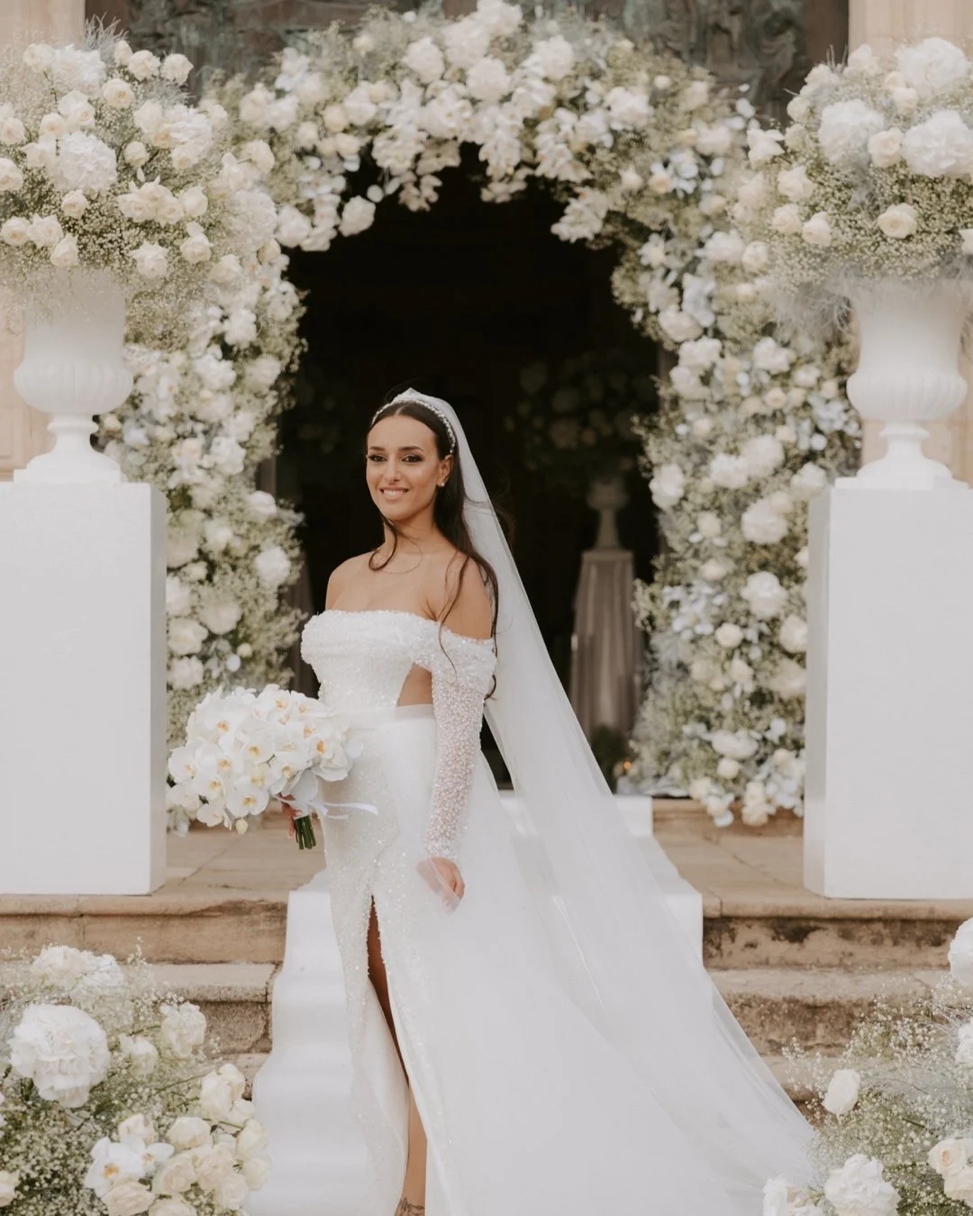 Foto di una sposa in abito bianco con bouquet di fiori bianchi, circondata da decorazioni floreali in bianco, in un'ambientazione elegante per un matrimonio.