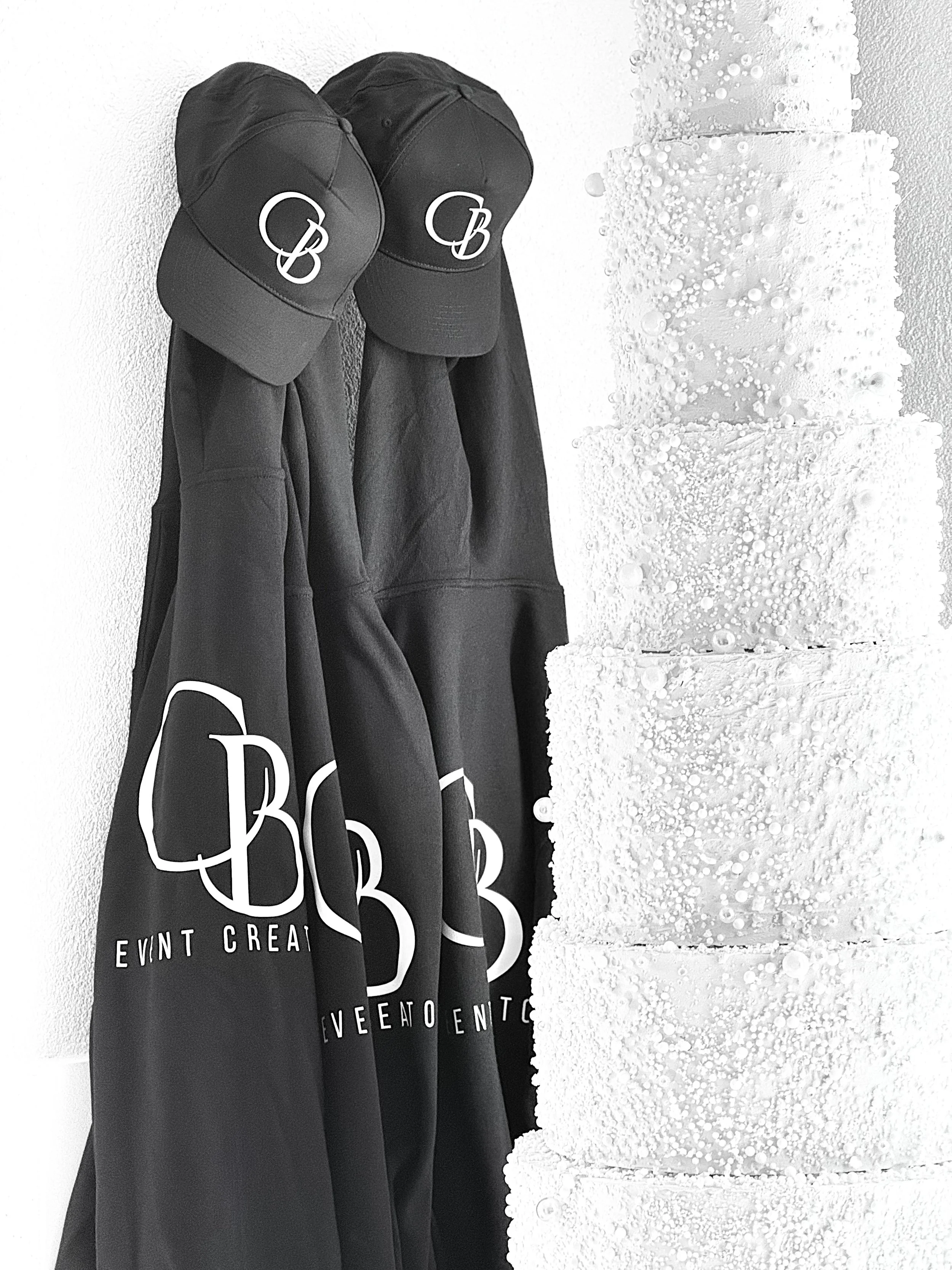 Tre cappelli neri appoggiati a un muro, con la scritta 'EVENT CREATION' in bianco e lettere decorative, accanto a una grande torta bianca decorata con palline e fiocchi.