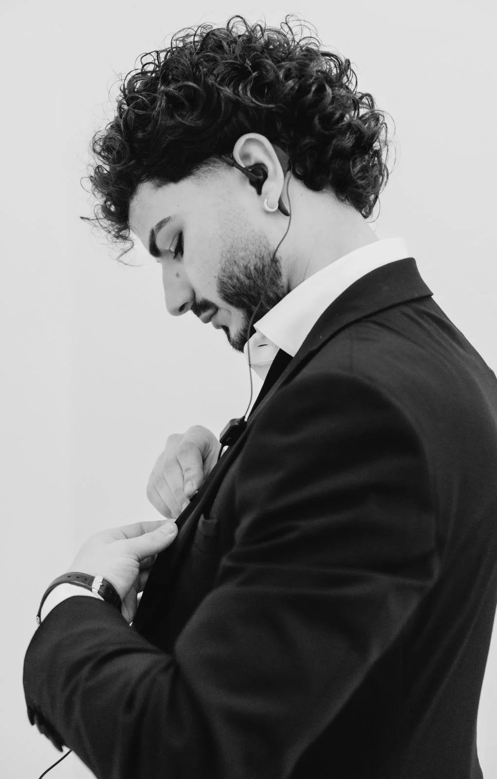 Ritratto in bianco e nero di un uomo con capelli ricci, barba e orecchino, mentre sistema la giacca, indossa una camicia bianca e una giacca scura, e ha cuffie auricolari.