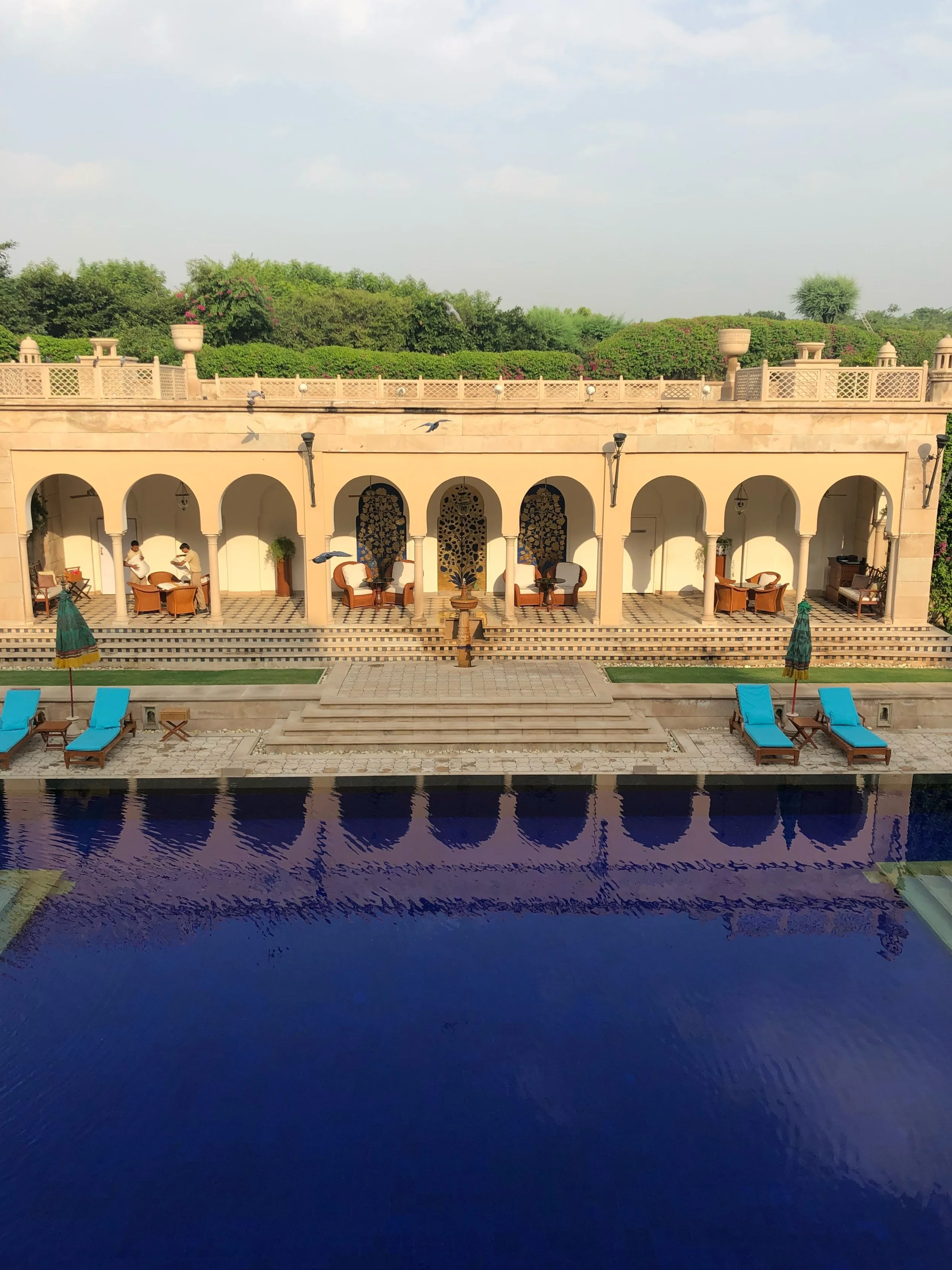 Luxurious outdoor swimming pool with blue water, surrounded by sun loungers and umbrellas, in front of an elegant building with arches, seating areas, and decorative panels, set against a backdrop of greenery and a clear sky.