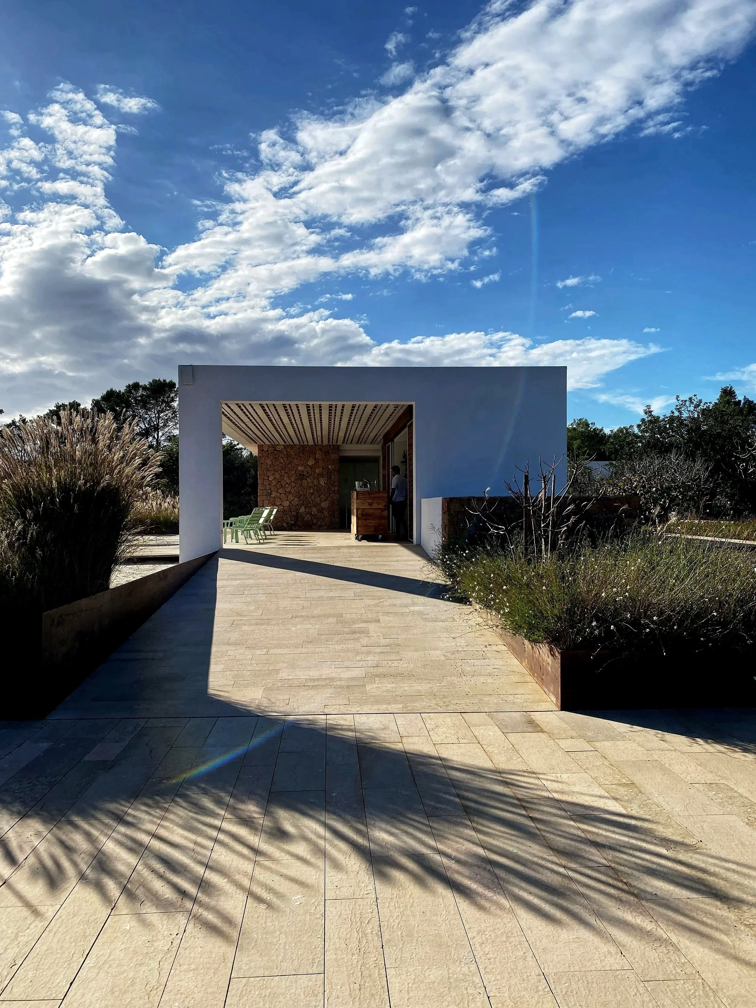 Modern building with a white facade and open entrance, surrounded by plants and trees under a partly cloudy sky.