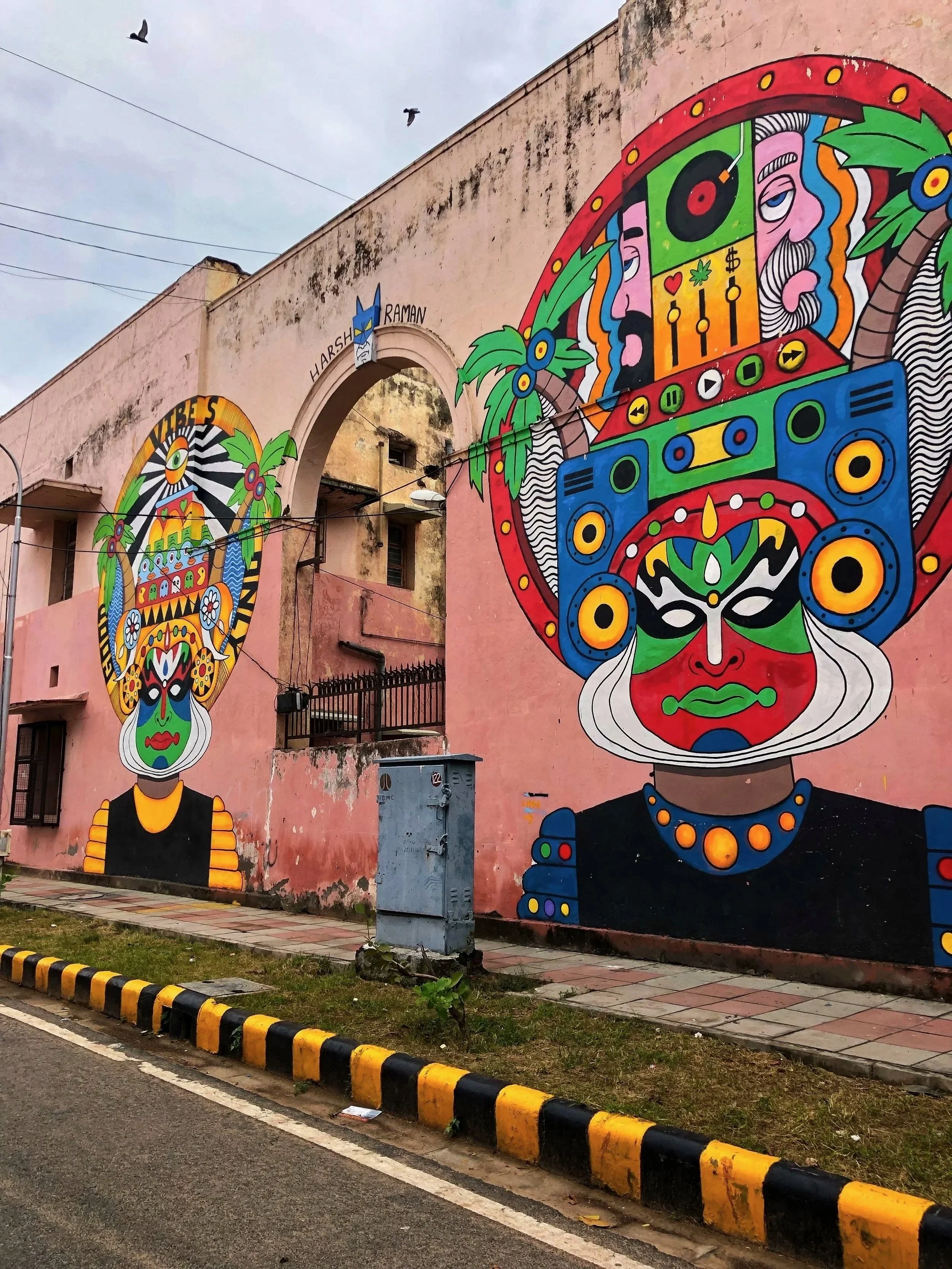 Colorful street mural featuring two faces with large, intricate headdresses decorated with flowers, vines, and abstract patterns. The mural is on the side of a pink building with an arched opening, and there are two birds flying in the sky above.