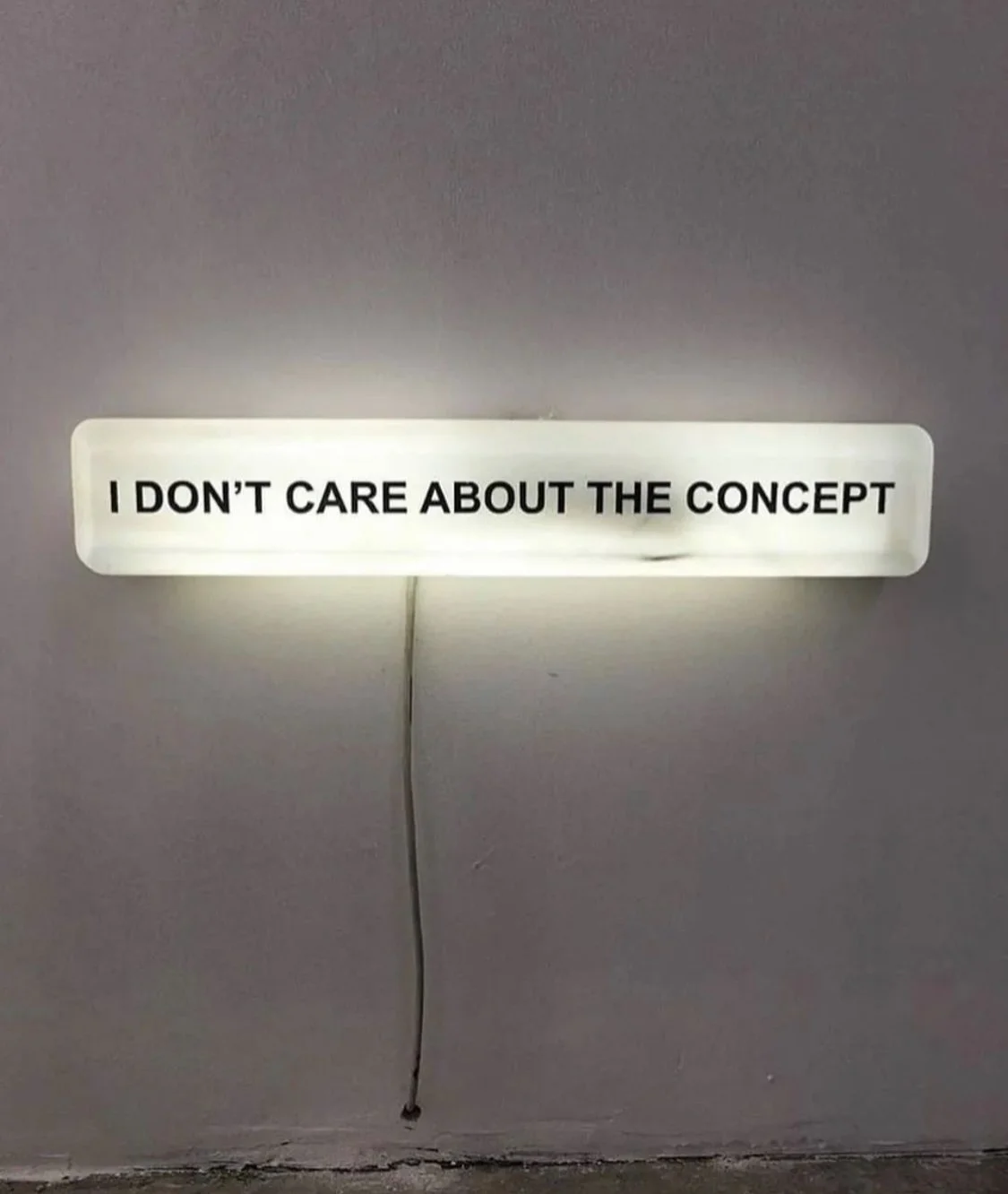 A white illuminated sign with black text reading "I DON'T CARE ABOUT THE CONCEPT" hangs on a plain wall with a visible power cord hanging beneath it.
