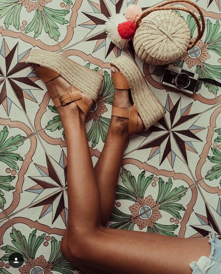 A person lying on a patterned tiled floor, wearing tan platform heels and denim shorts. Items around include a vintage camera, a straw bag, and a pom-pom hair accessory with red, white, and pink pom-poms.