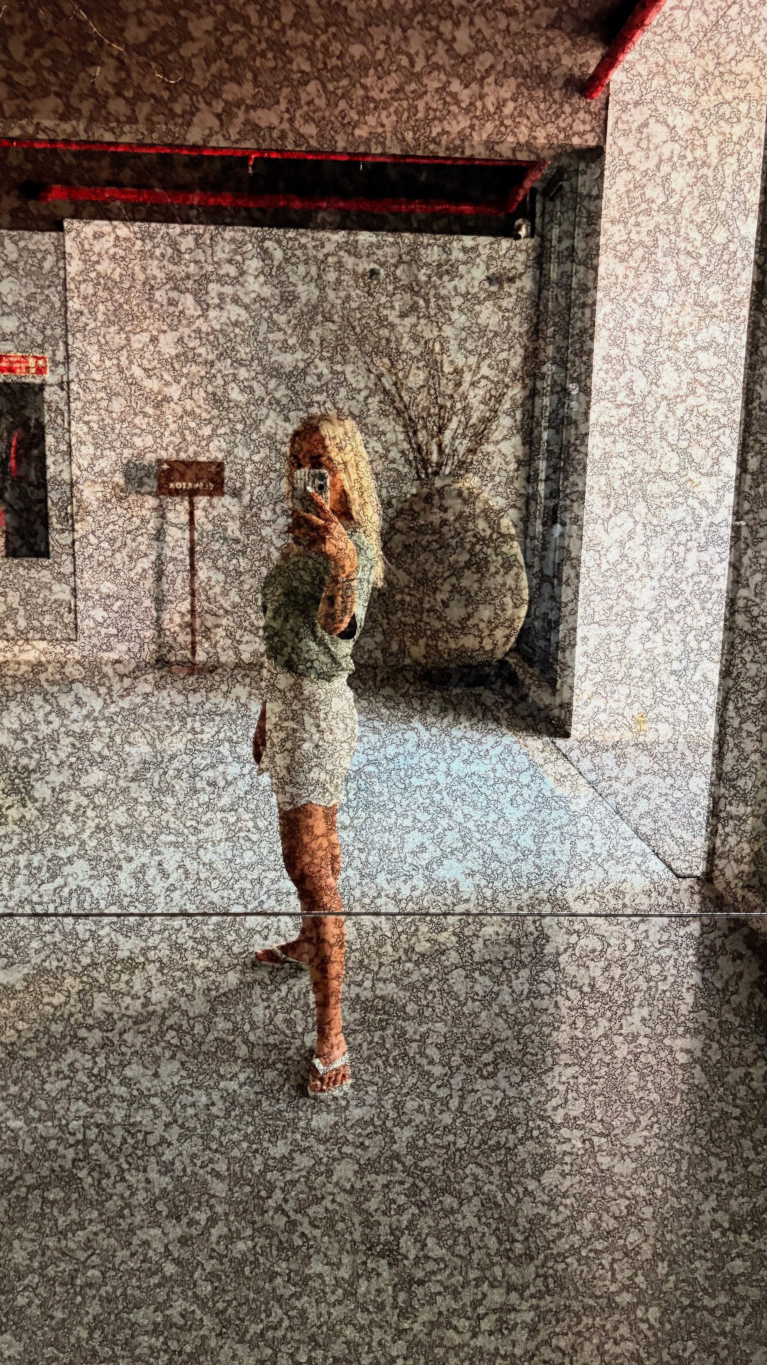 A woman in a white dress taking a mirror selfie in a room with patterned walls and floor, with a black and white artwork or bulletin on the wall and a large decorative branch behind her.