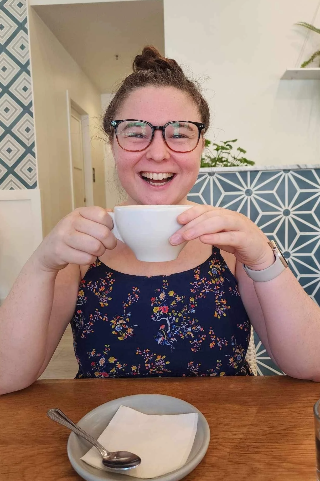 Co-Founder Cathlyn Melvin, wearing glasses and a blue flowered dress, drinks a mug of tea and smiles at the camera - Merry Berry Confection Co, a micro-bakery specializing in holiday bakes and dessert bars for weddings and events near Raleigh, NC