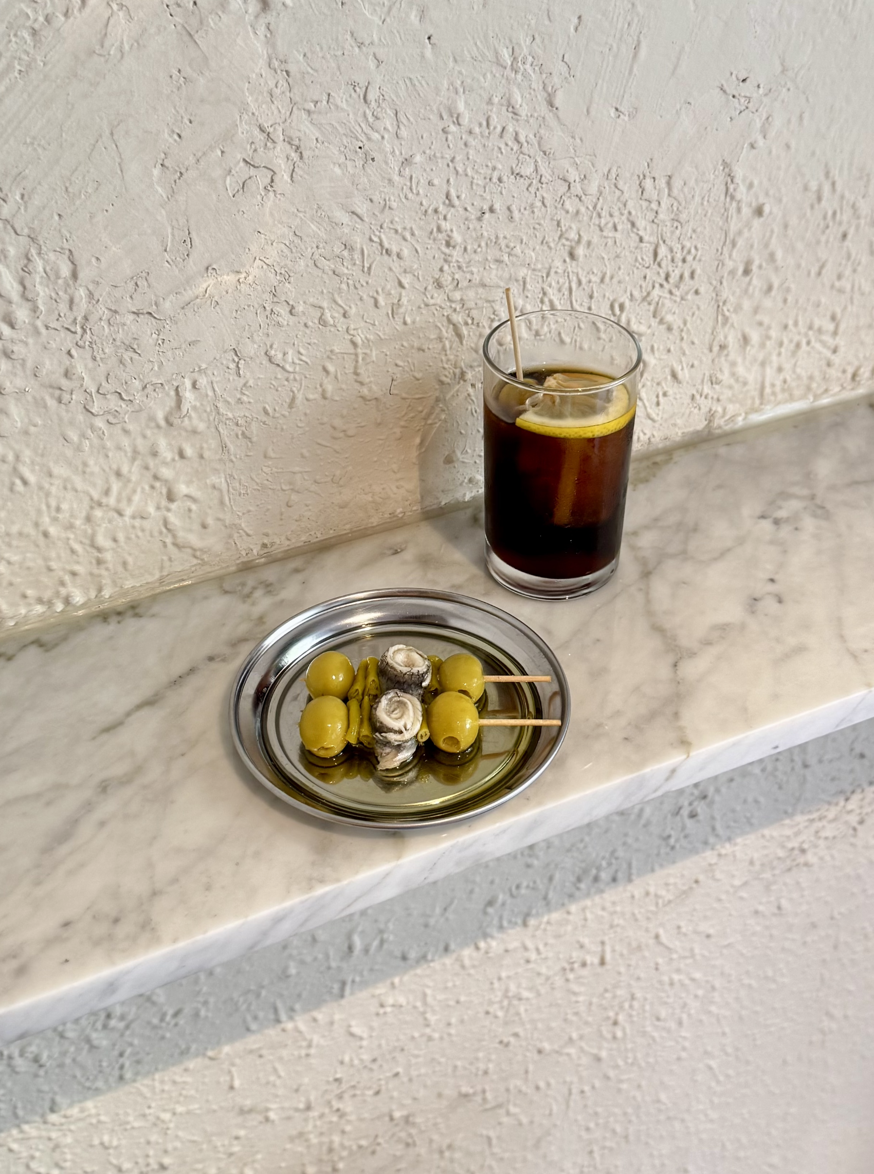 Un vaso de refresco con limón y una bandeja con aceitunas y boquerones