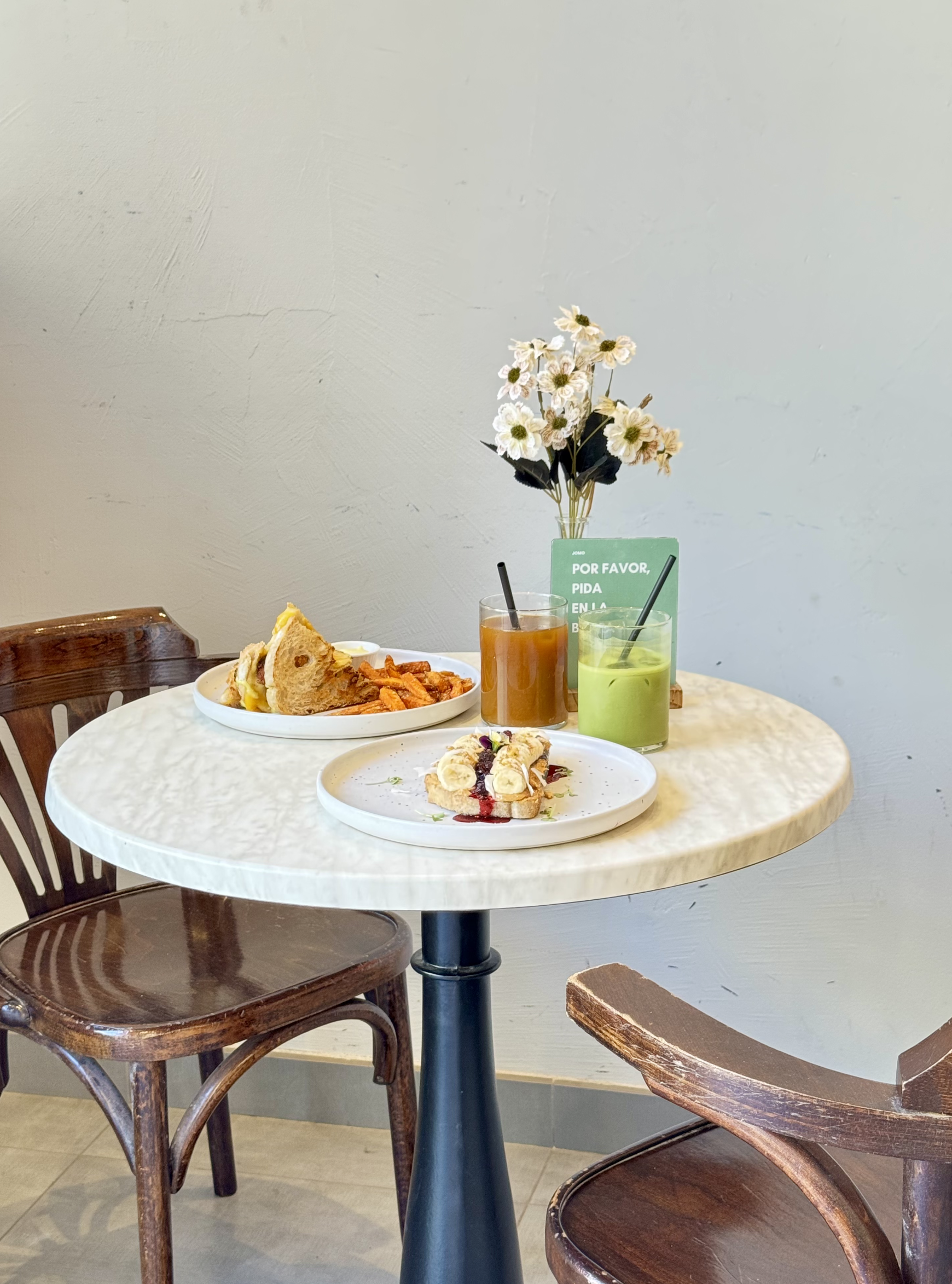 Mesa con dos platos de comida, dos bebidas y flores en un florero.