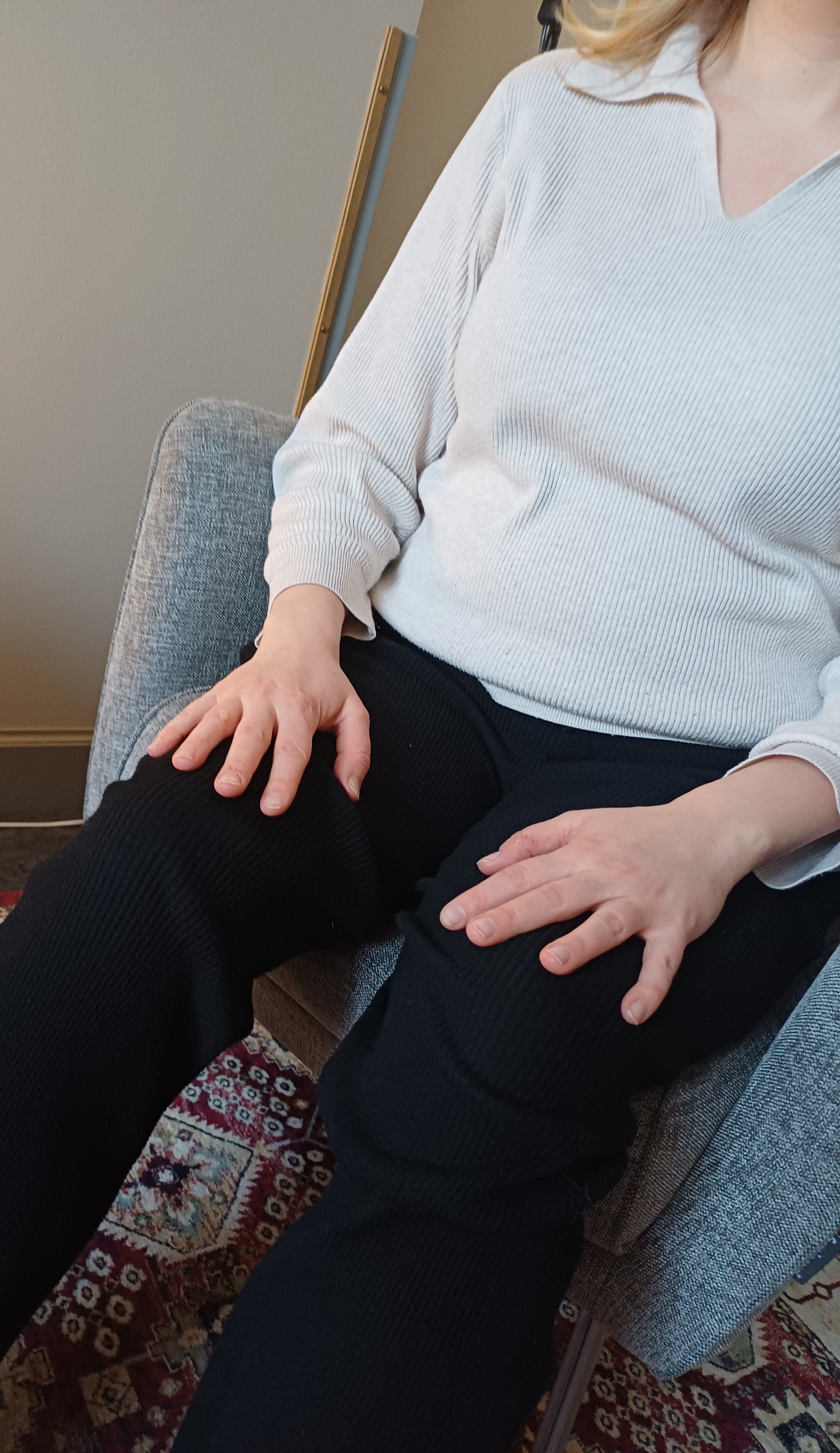 A woman sitting on a gray upholstered chair, wearing a white ribbed sweater and black pants, resting one hand on her thigh and the other on her knee, with a patterned rug beneath the chair.
