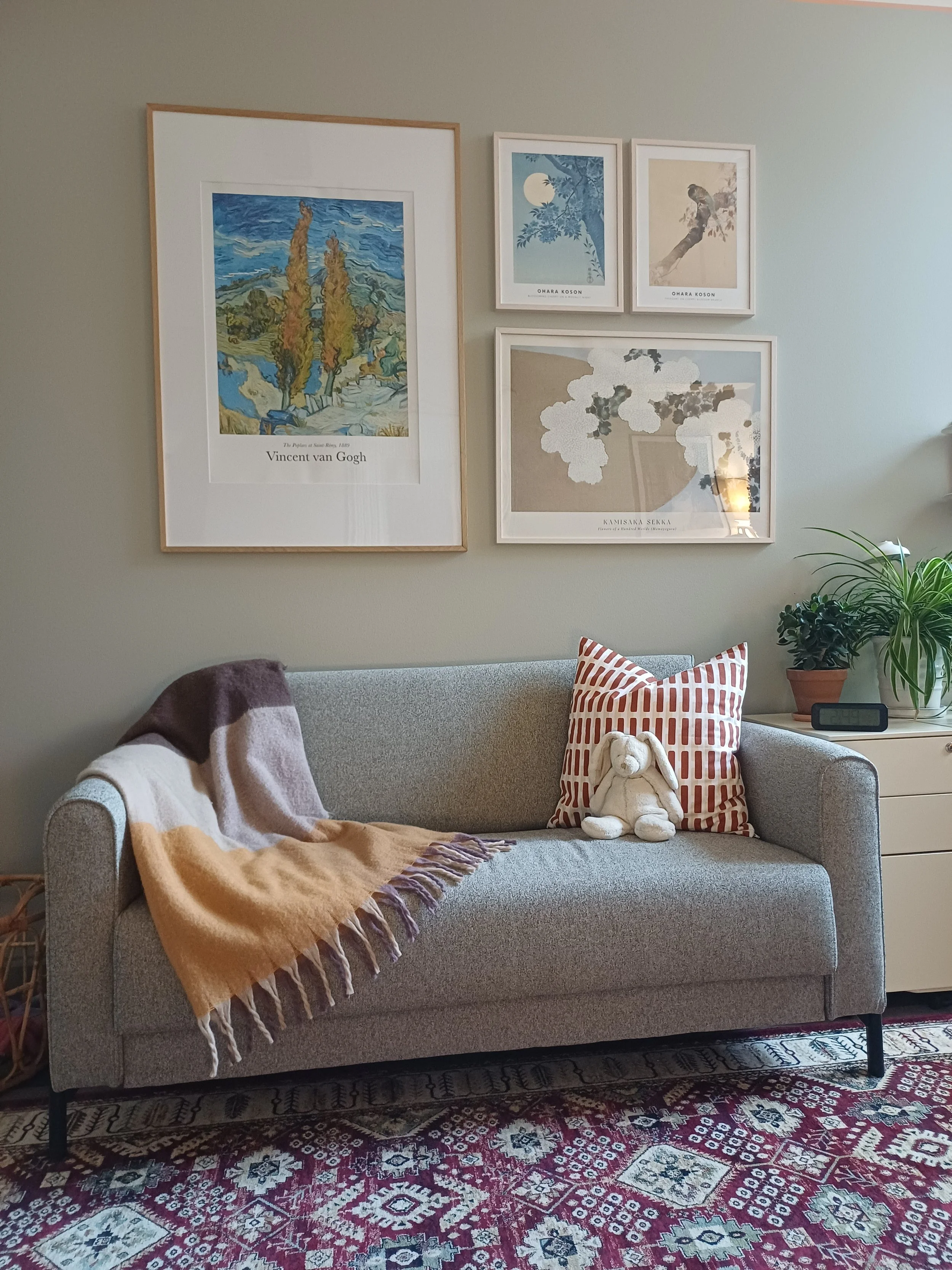 Interior wall with five framed art prints, including a Vincent van Gogh poster, and a wooden cabinet with plants, a vase with dried flowers, candles, a clock, and a desk lamp.
