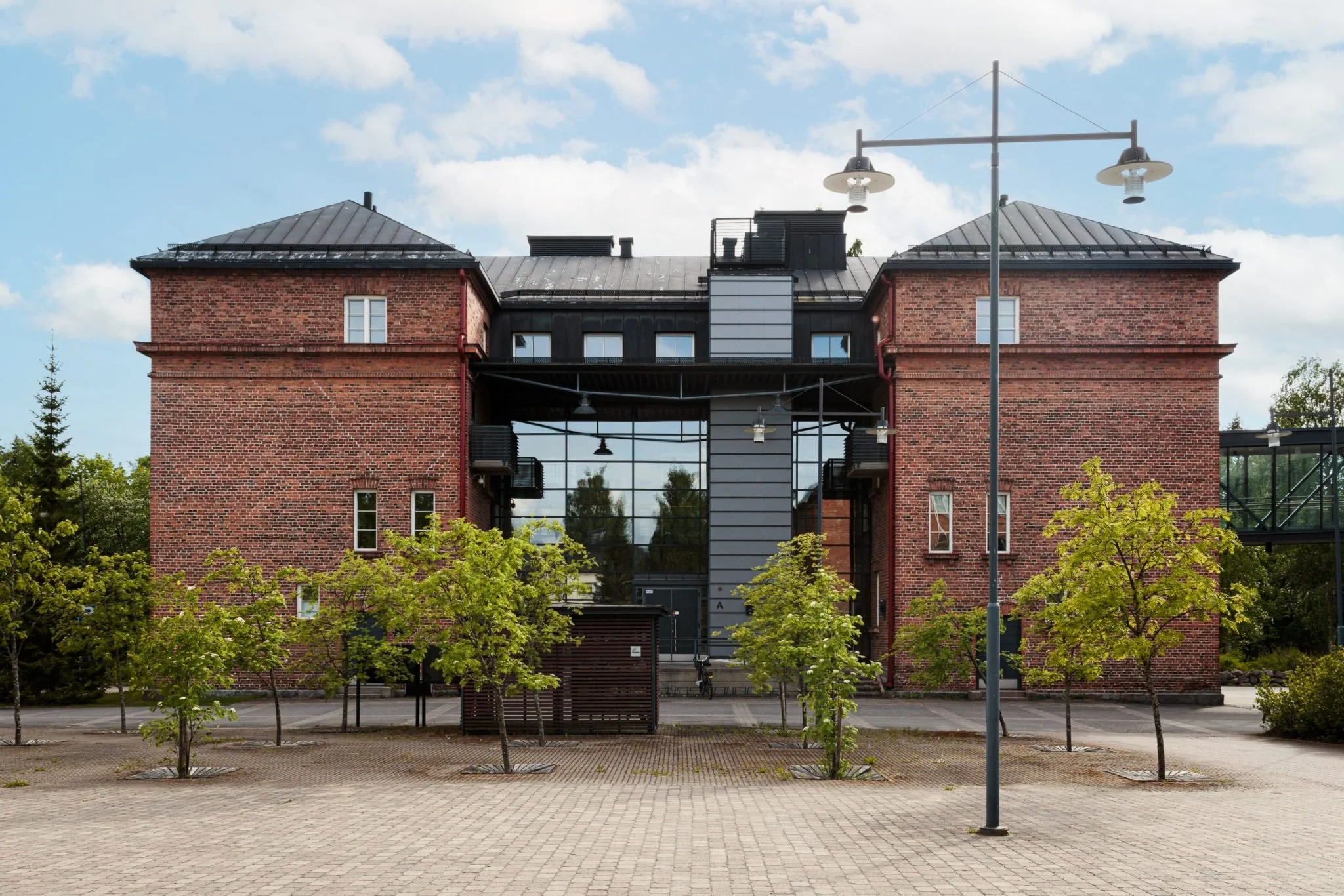 A modern brick building with large glass windows, situated behind small trees and a paved plaza, under a partly cloudy sky.