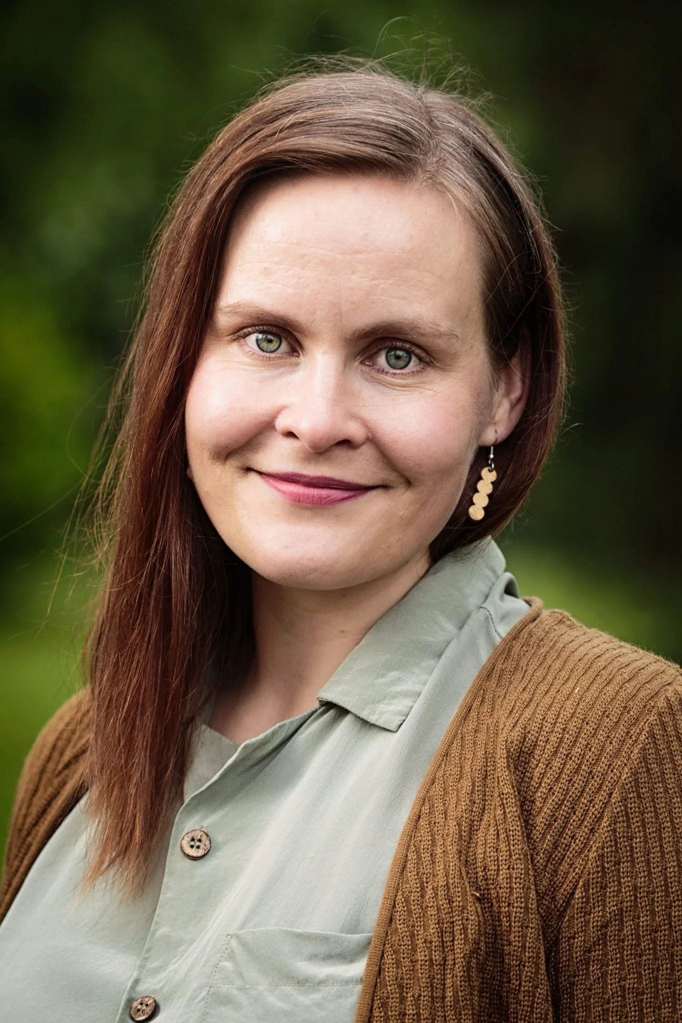 Close-up portrait of a woman with shoulder-length brown hair, light-colored eyes, wearing a light green shirt and a brown cardigan, outdoors with a blurred green background.