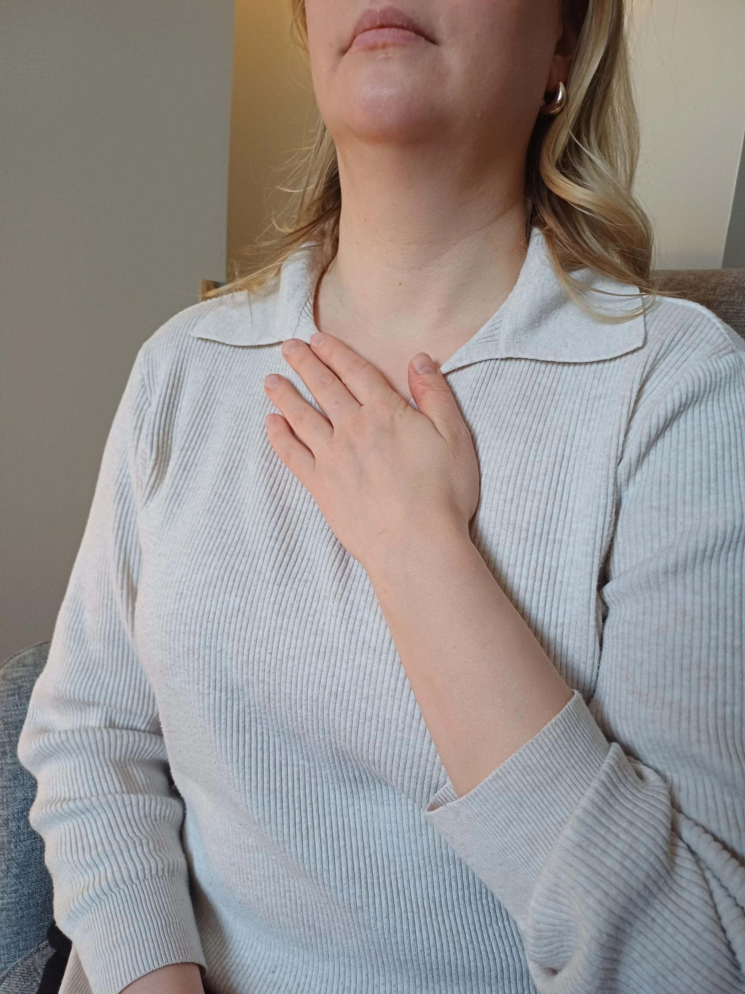 A woman with blonde hair wearing a light beige collared shirt is seated indoors with her hand on her neck. The background includes neutral-colored walls and furniture.