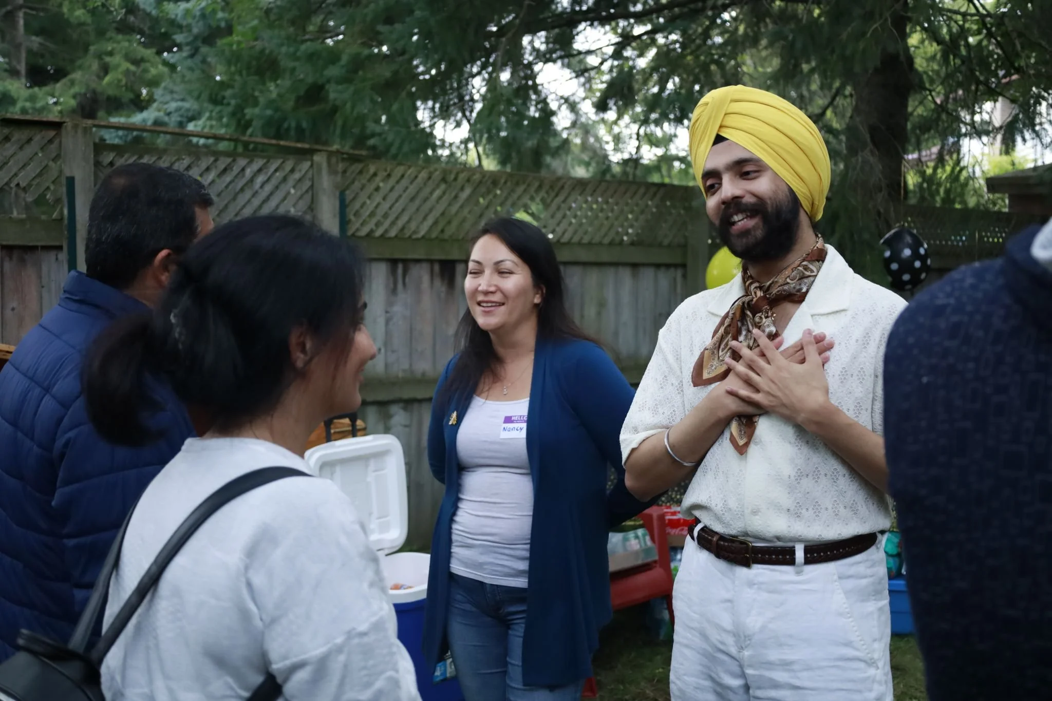 People having a conversation at an outdoor social gathering, with a man wearing a yellow turban and a white shirt, and women in casual attire, in a backyard with Christmas balloons and a wooden fence.