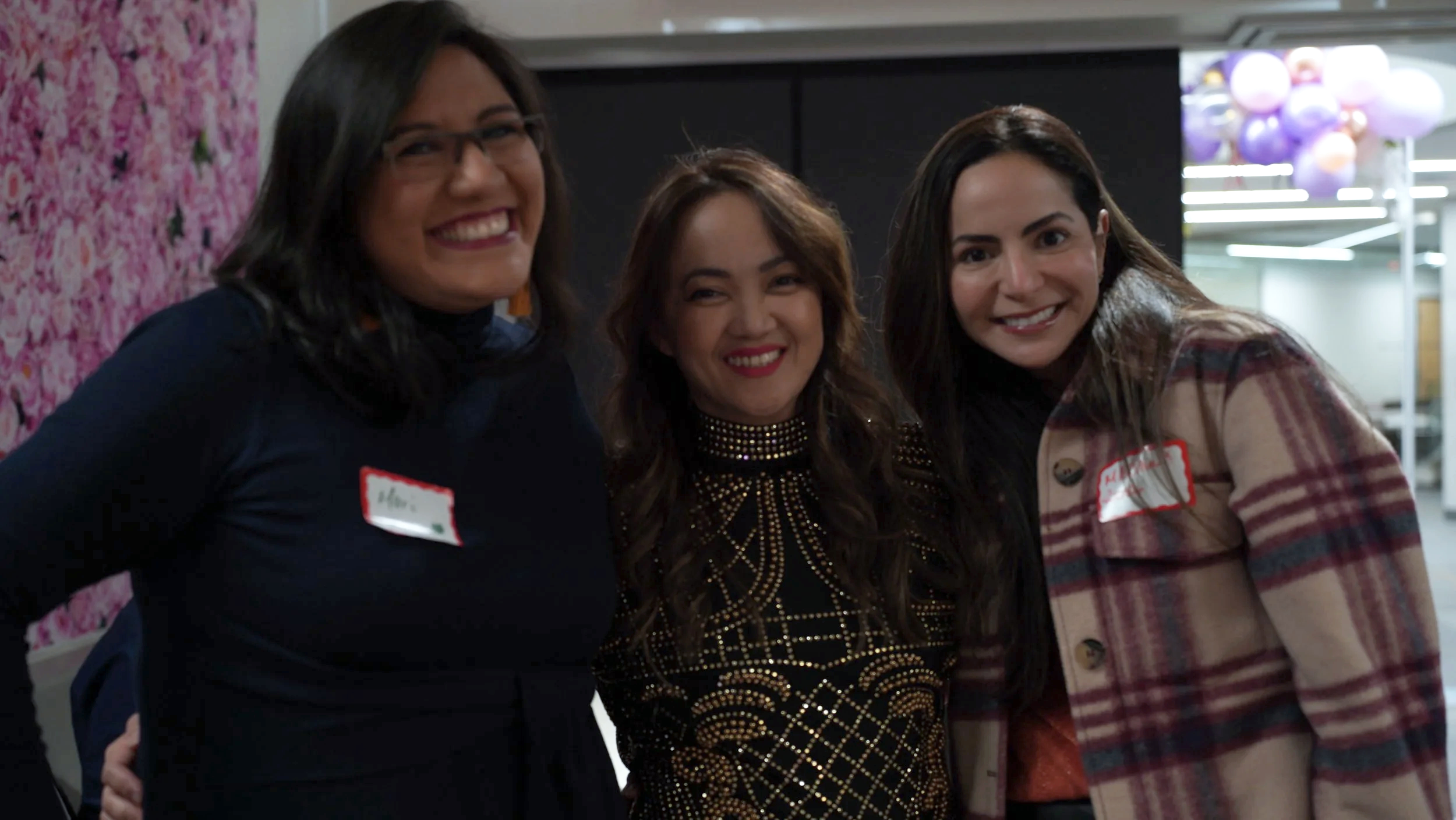 Three women smiling at an indoor event, wearing name tags and casual outfits.