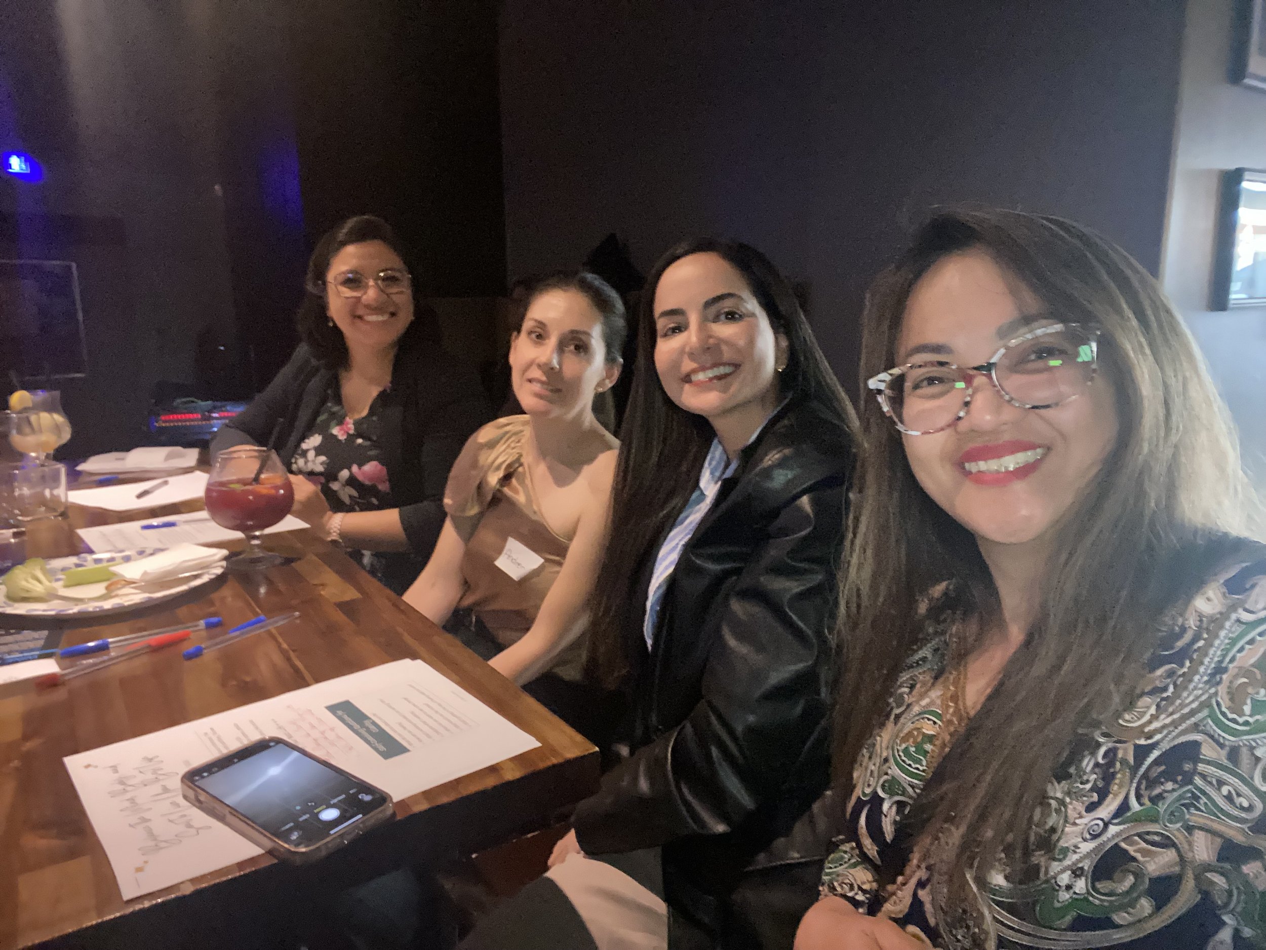 Four women smiling and sitting at a bar or restaurant table with drinks and papers, with a wireless microphone and colorful pens on the table.