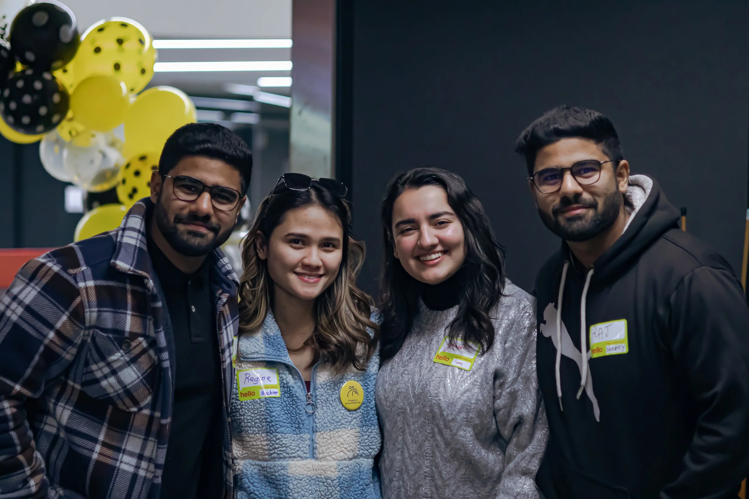 Group of four young adults smiling at a social event, with yellow and black balloons in the background.