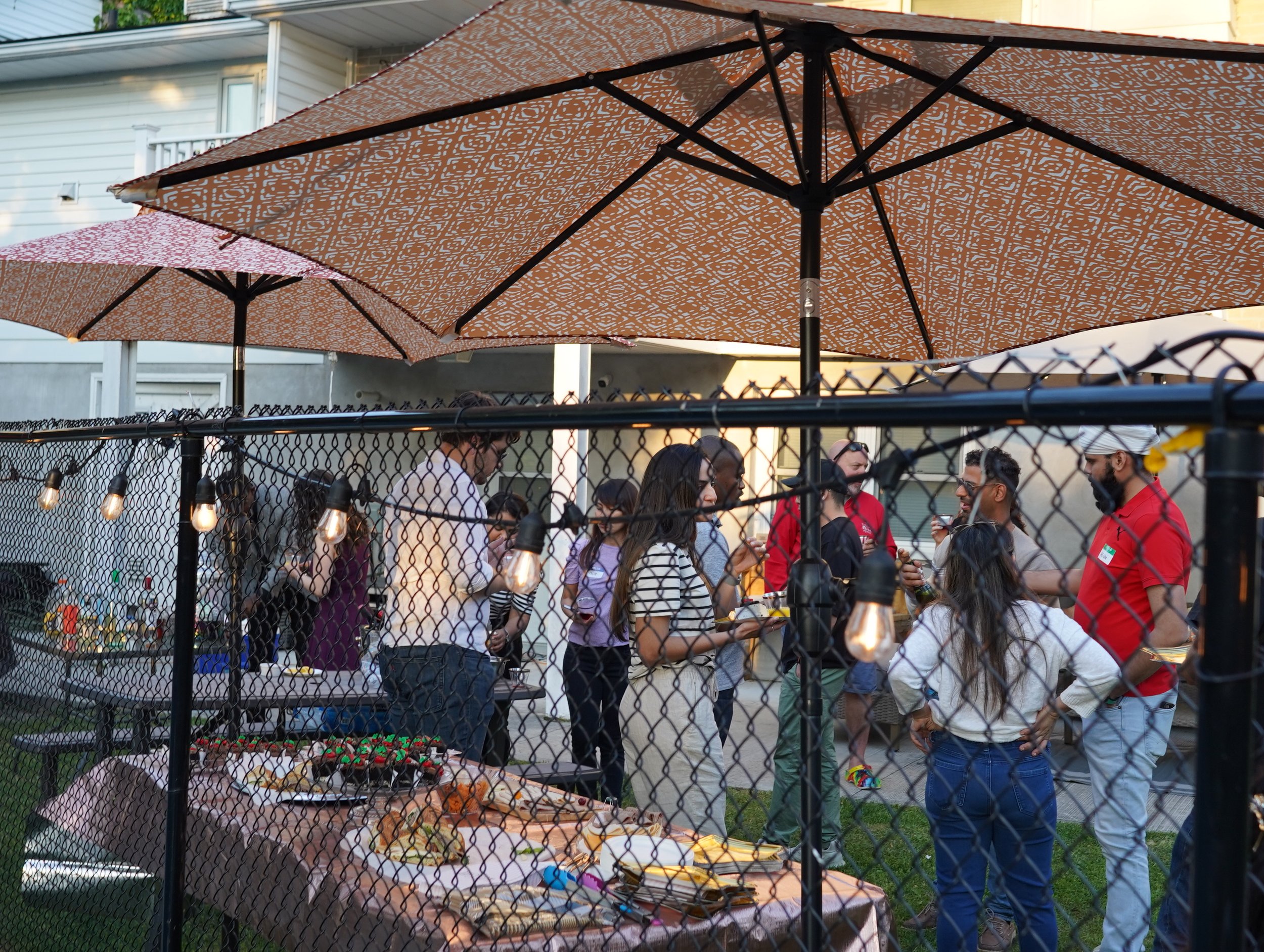 People gathered at an outdoor party or gathering, through a black chain-link fence with string lights, with large umbrellas providing shade, and a table with food and snacks in the foreground.