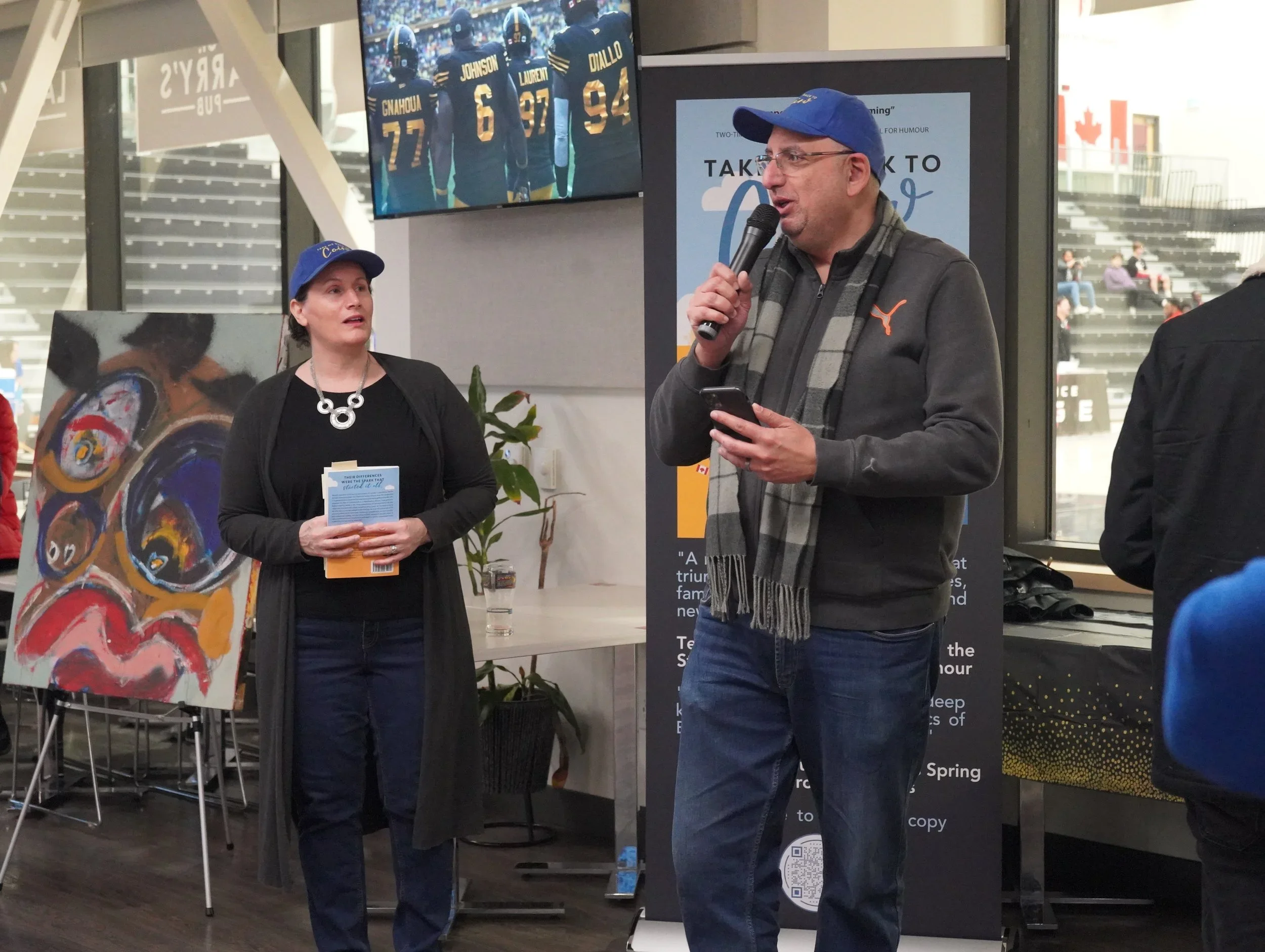 A man speaking into a microphone while a woman looks on, with a television showing football players in the background and an abstract painting on an easel beside her.