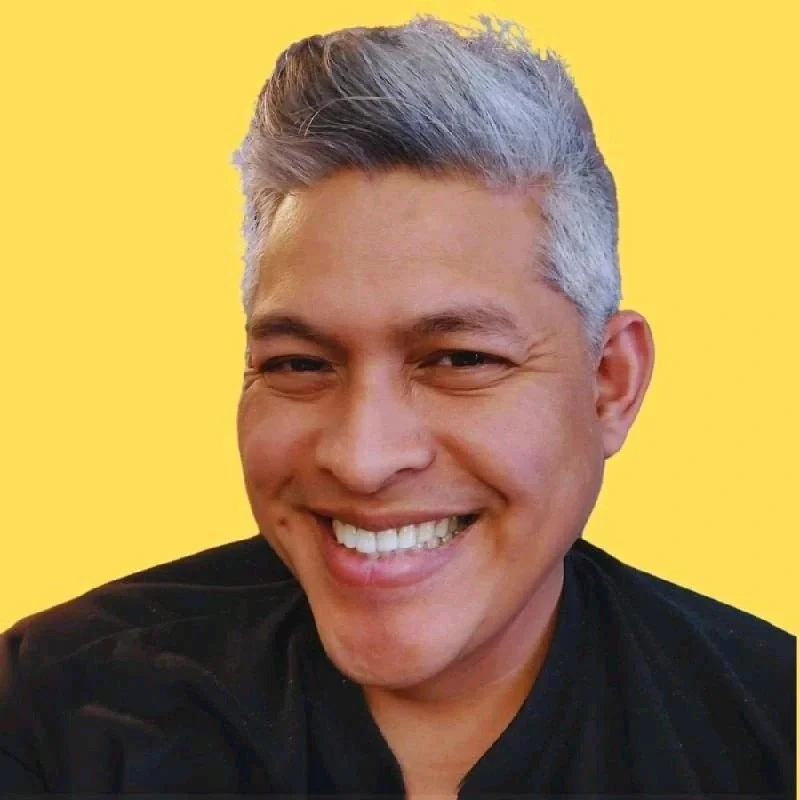 A smiling man with gray hair and wearing a black shirt, against a yellow background.