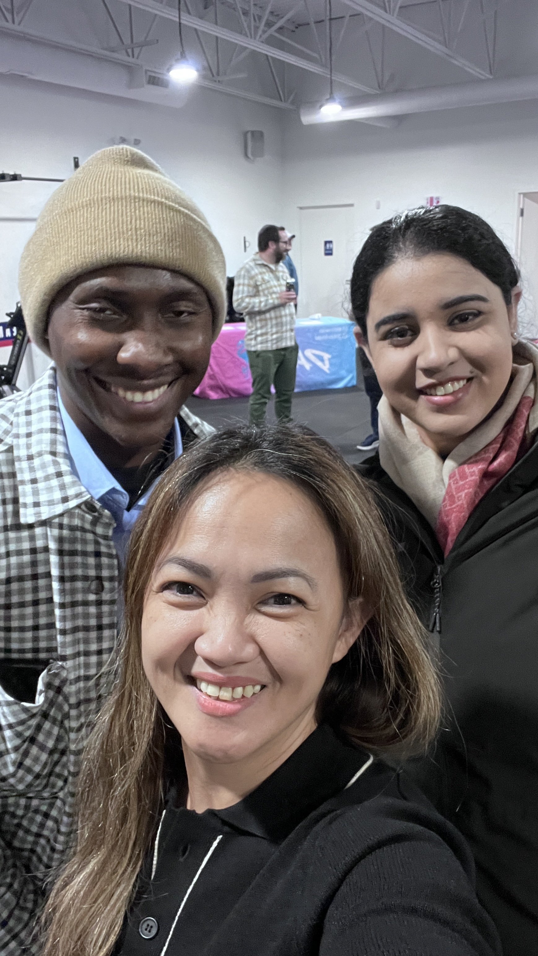 A group selfie featuring three smiling people in a room with a white ceiling and walls, with a guy wearing a beige beanie and checkered shirt, a woman with dark brown hair in black top, and another woman with black hair and a beige scarf. In the background, there are people and colorful tables.